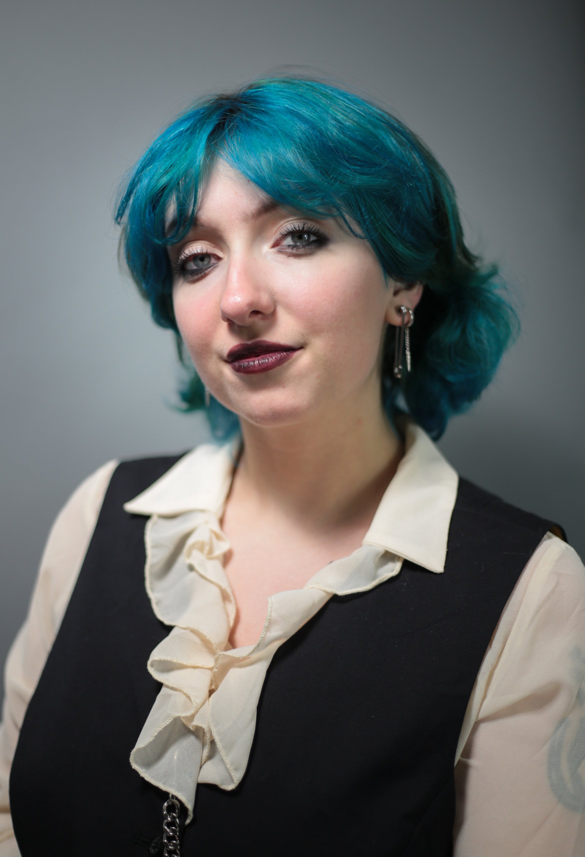 A woman with short, layered blue hair and light eyes, wearing dark lipstick, earrings, a cream ruffled blouse, and a black vest, standing against a plain gray background.
