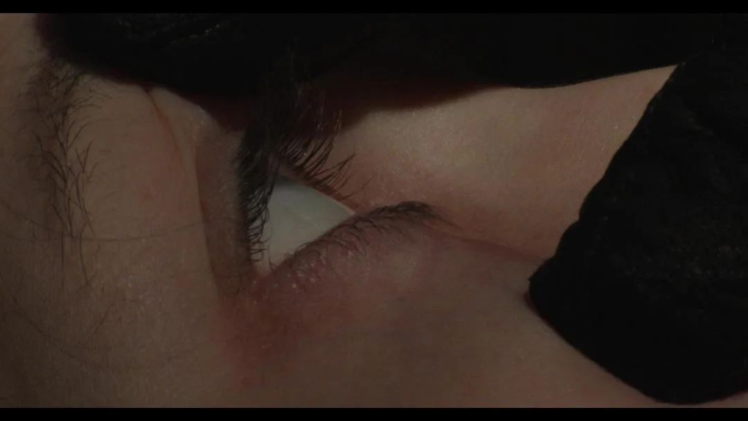 Close-up of a person's eye with makeup, visible eyelashes and skin texture, with a black cloth or garment partially covering the face.