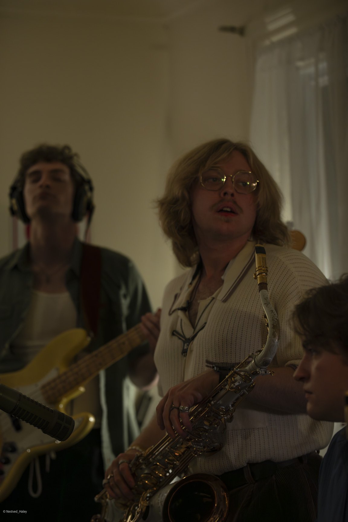 Three men in a music studio, one playing a guitar, another with a saxophone, and the third looking on.