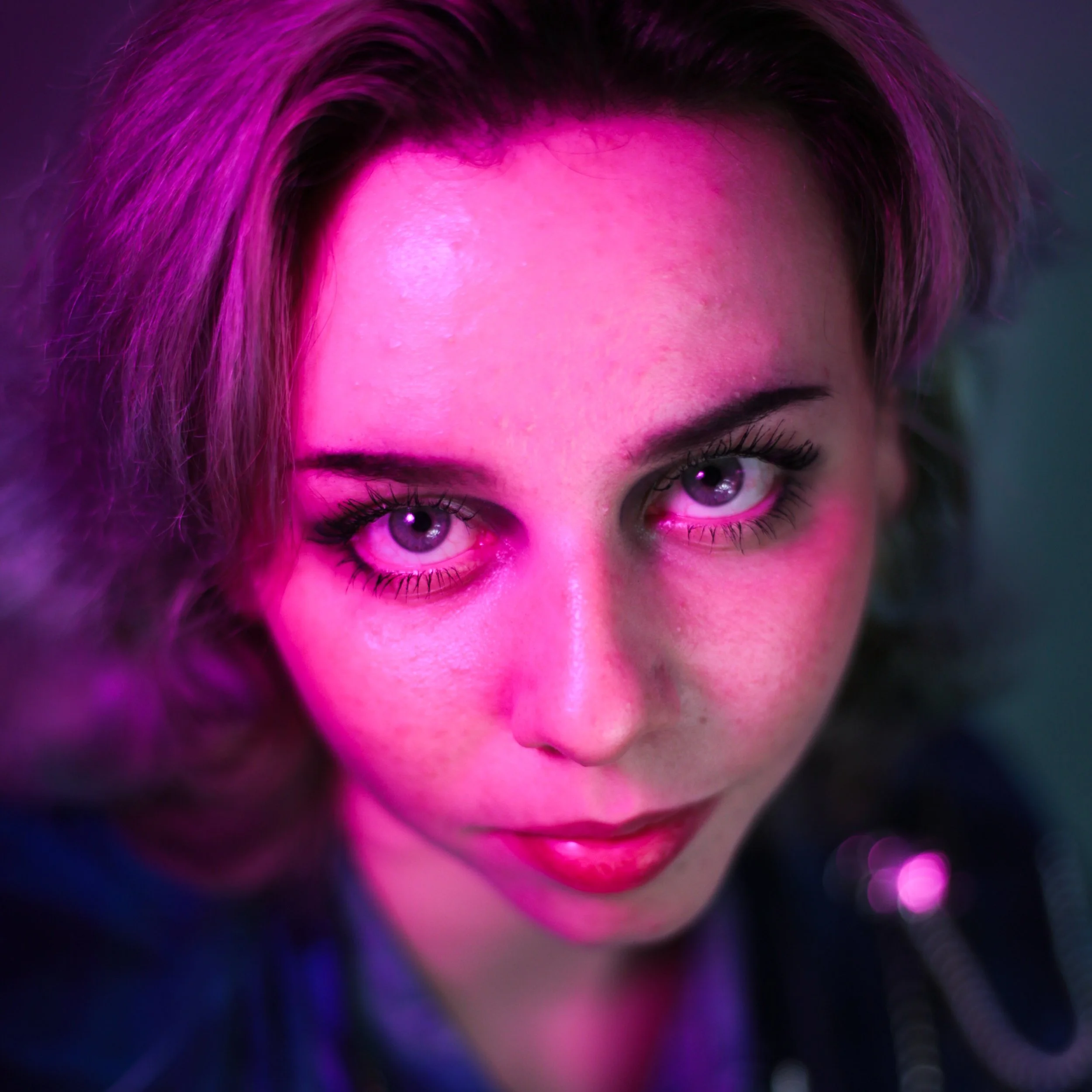 Close-up of a woman with purple eyes and short, mullet-style hair, illuminated by pink and purple lighting.