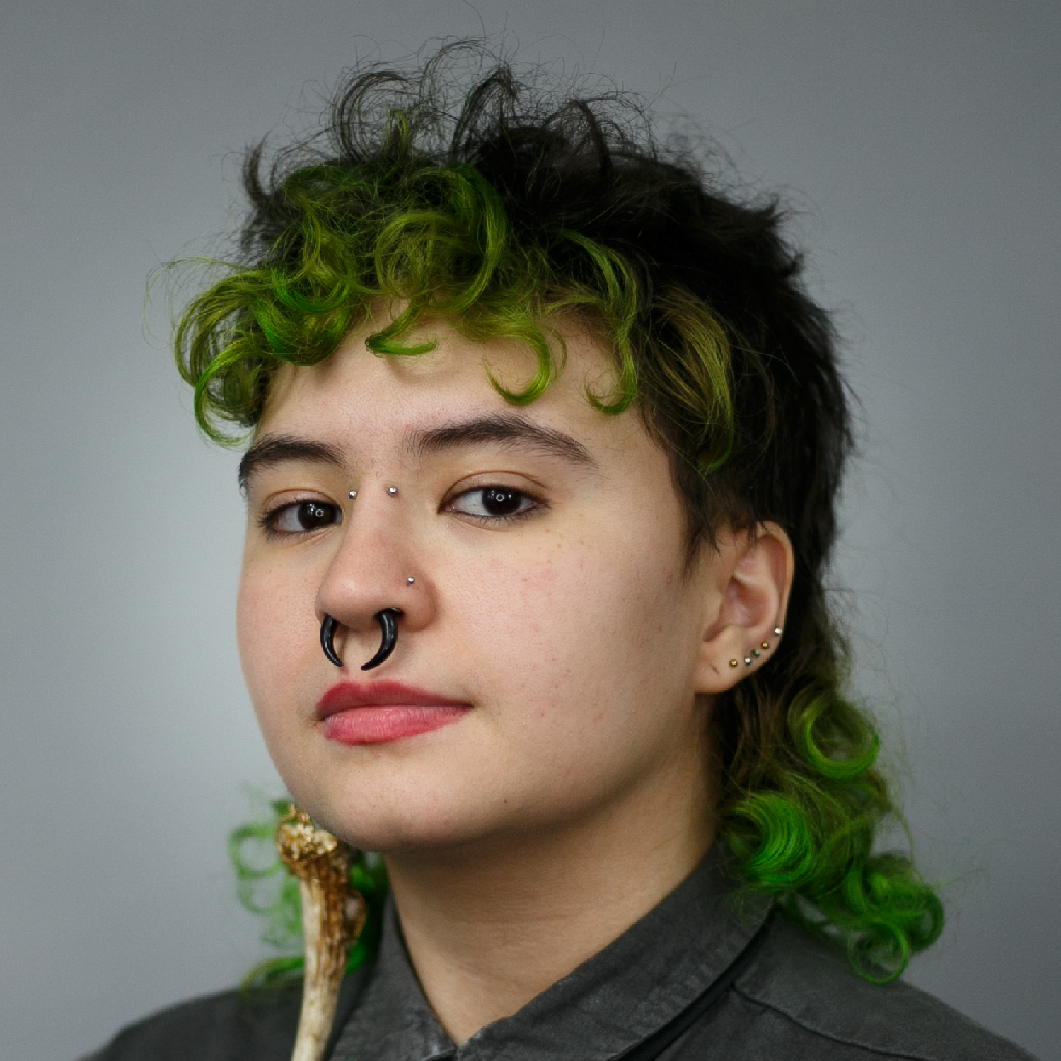 Person with green-dyed curly hair, multiple facial piercings, and dark clothing, with a pipe in their mouth.