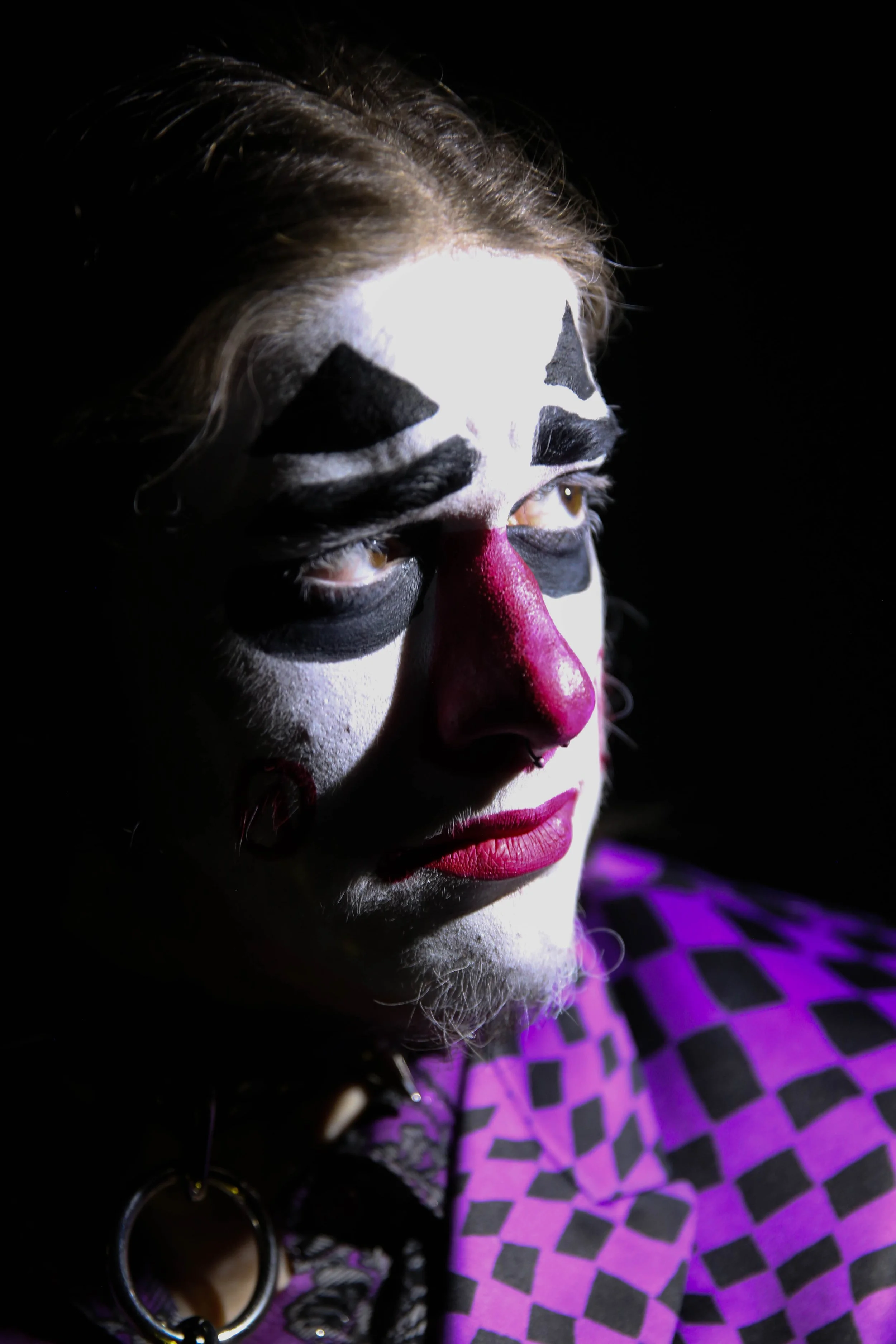 Person with clown makeup featuring black and white face paint, a red nose, and pink lipstick, wearing a purple and black checkered garment.