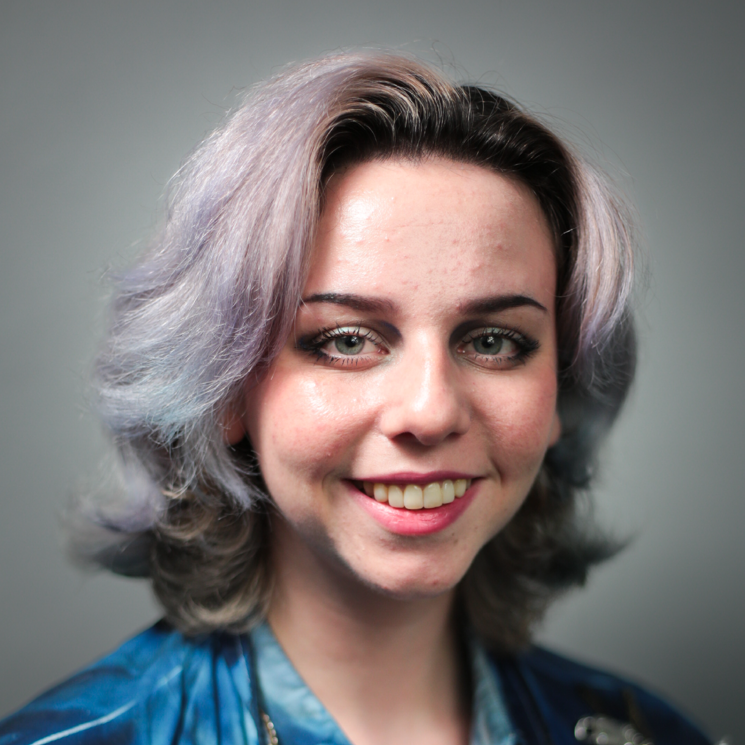Close-up portrait of a young person with ash blonde hair, green eyes, and a bright smile, wearing a blue top against a neutral background.