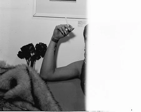 Close-up black and white photo of a person with a tattoo on their arm holding a cigarette, with fur and dark foliage in the foreground.