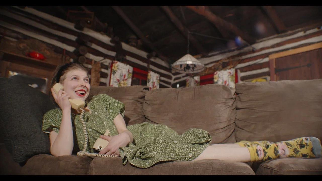 A woman in a vintage green dress with floral pattern, colorful socks, and makeup, lying on a brown couch in a rustic log cabin interior, talking on a rotary phone, smiling happily.