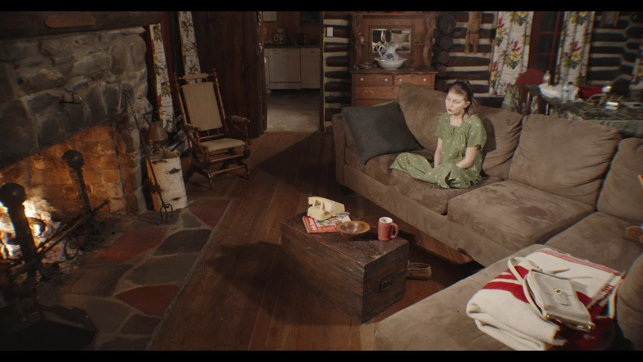 A woman sitting alone on a brown couch in a cozy living room near a lit fireplace. The room has wooden walls, a rocking chair, a wooden chest used as a coffee table with a telephone, a plate, and a mug on it. There are curtains with floral patterns a