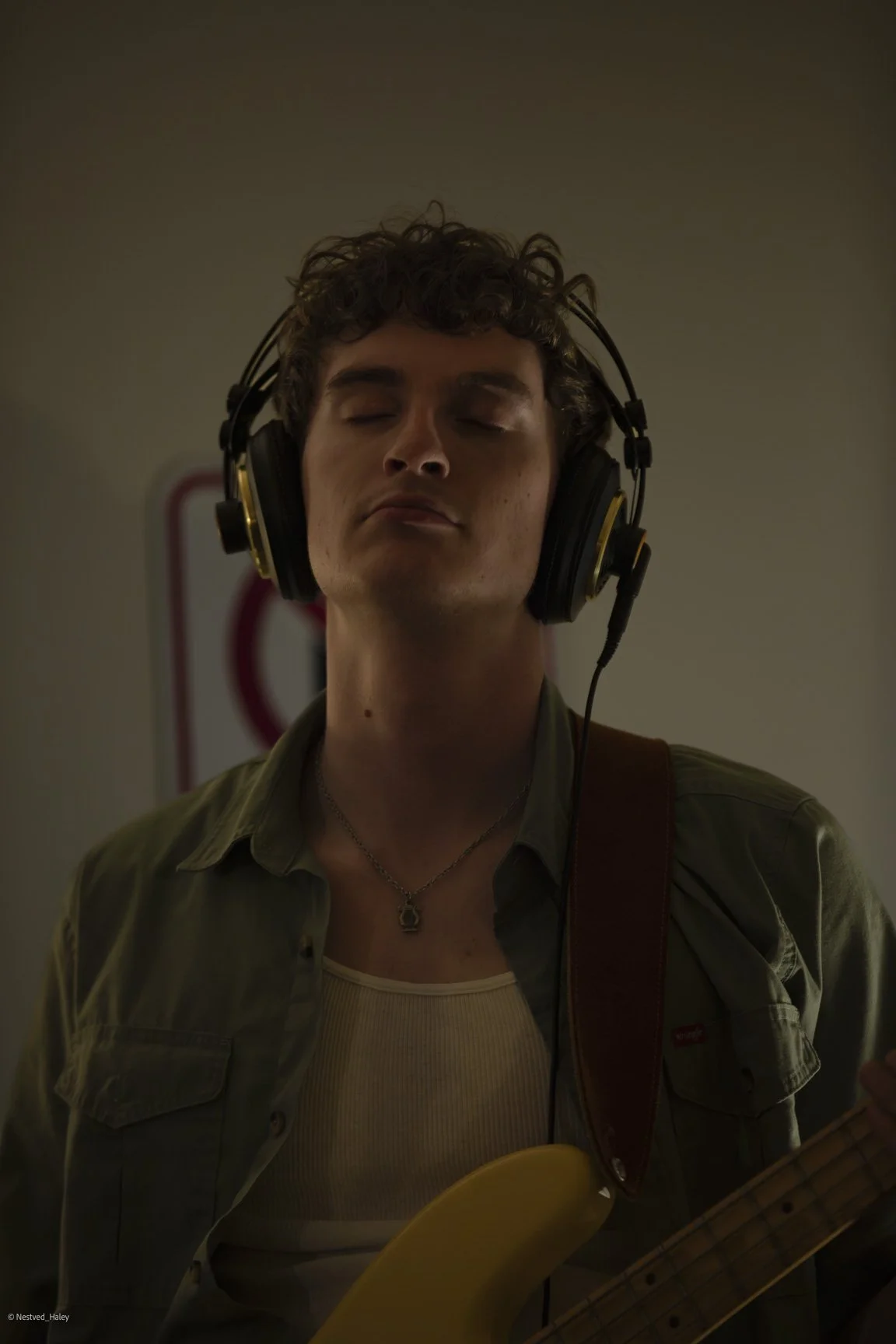Young man with curly hair wearing large over-ear headphones, closed eyes, holding a yellow electric bass guitar, wearing a green jacket and a white tank top, in a dimly lit room.