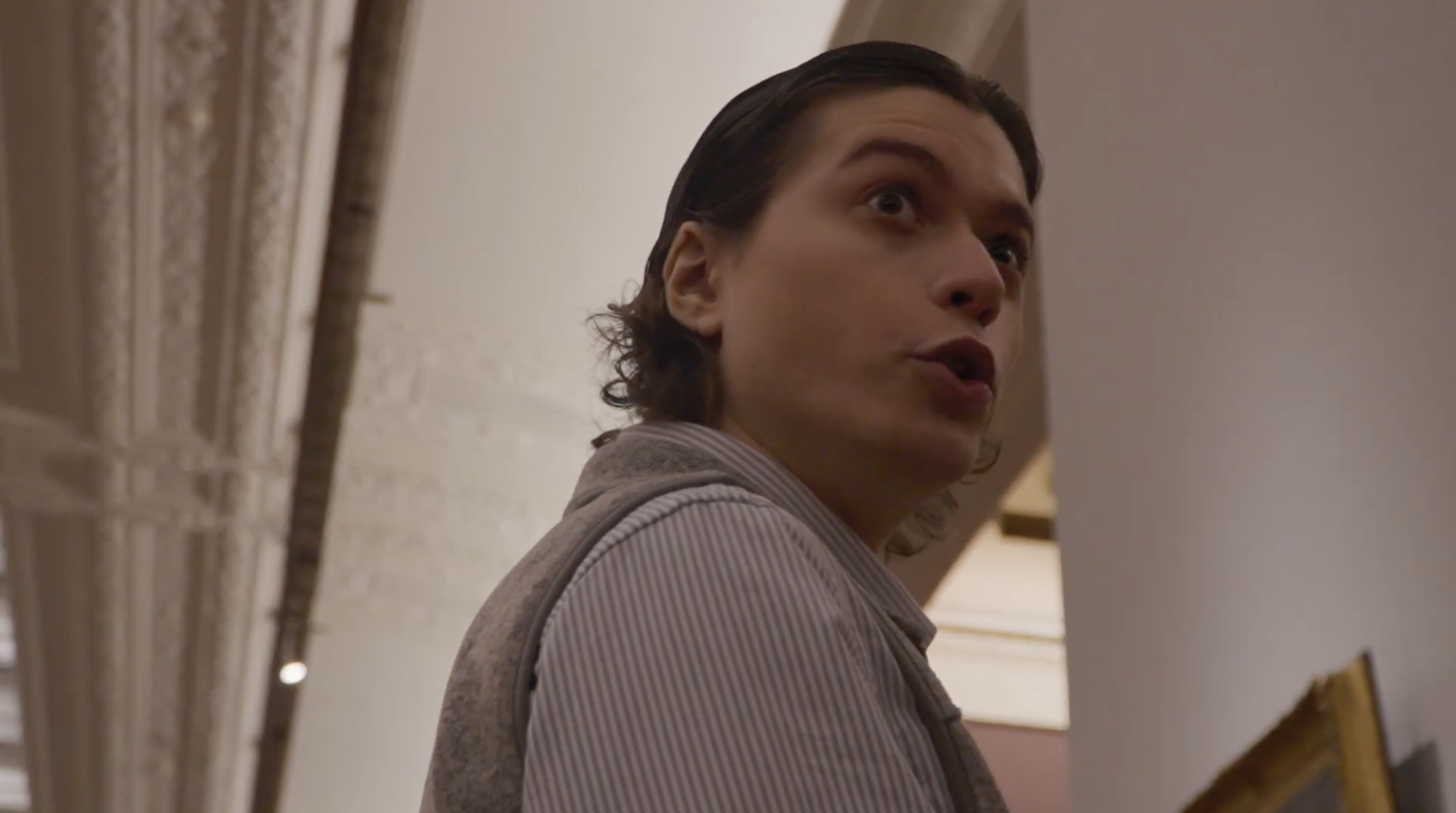 A person with dark, shoulder-length hair and wearing a striped shirt appears to be speaking, with their mouth open and eyes wide, standing indoors near a wall with ornate molding.
