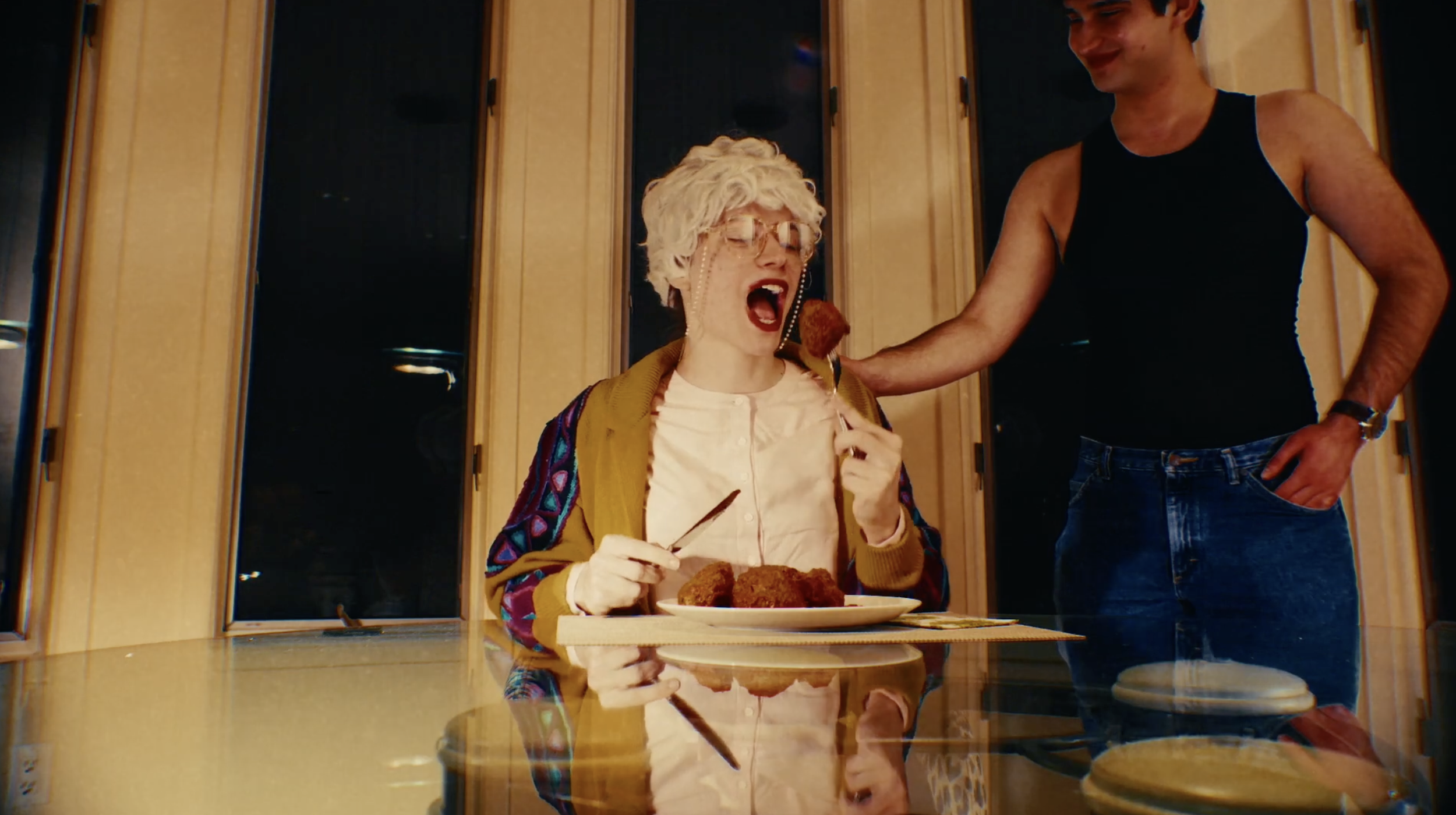 A woman with gray curly hair, glasses, and a colorful jacket is sitting at a dining table with food on her plate. A man, dressed in a sleeveless black shirt and jeans, is standing next to her offering her a piece of food on a fork.