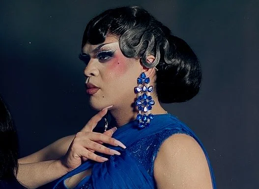 A person with dark hair styled in vintage waves, heavy makeup including blue eyeshadow and blush, wearing large blue floral earrings and a matching blue dress, with a hand gently touching their face.