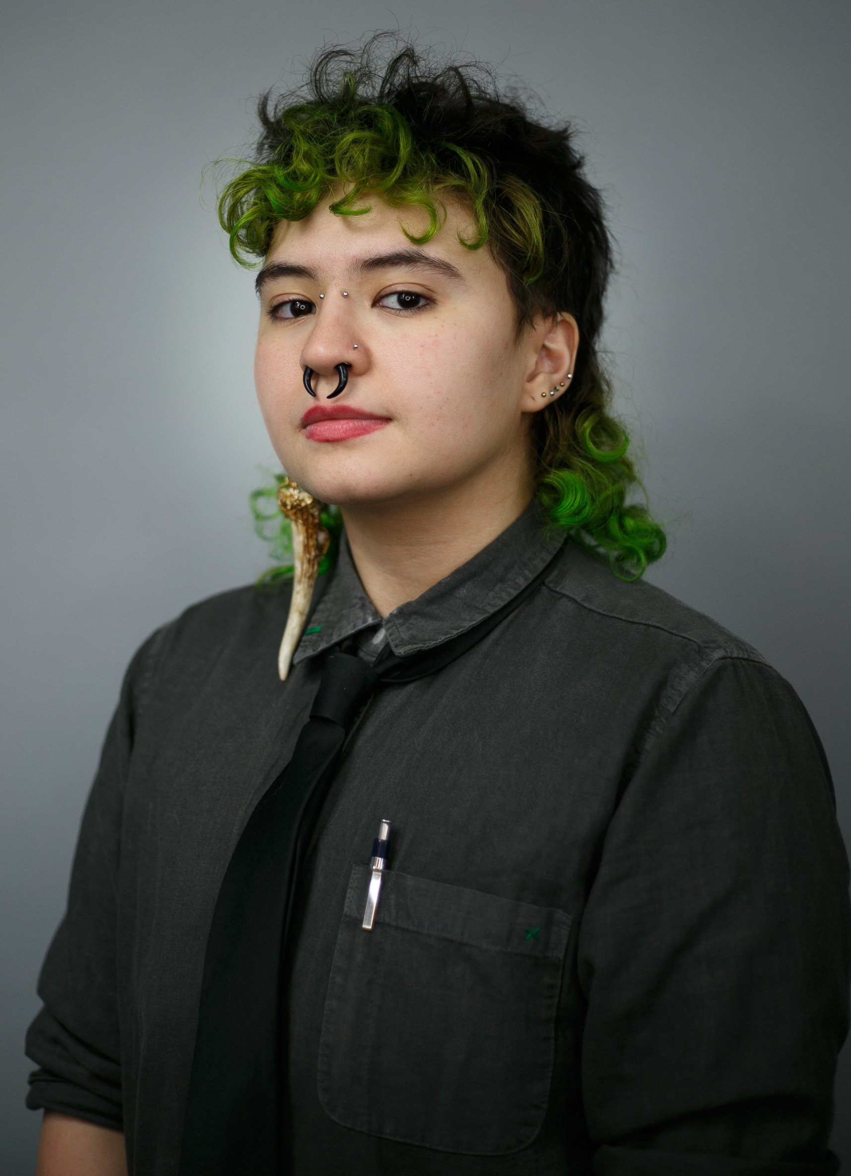 Person with green curly hair and multiple facial piercings wearing a dark shirt and tie, holding a horn-shaped object in their mouth.