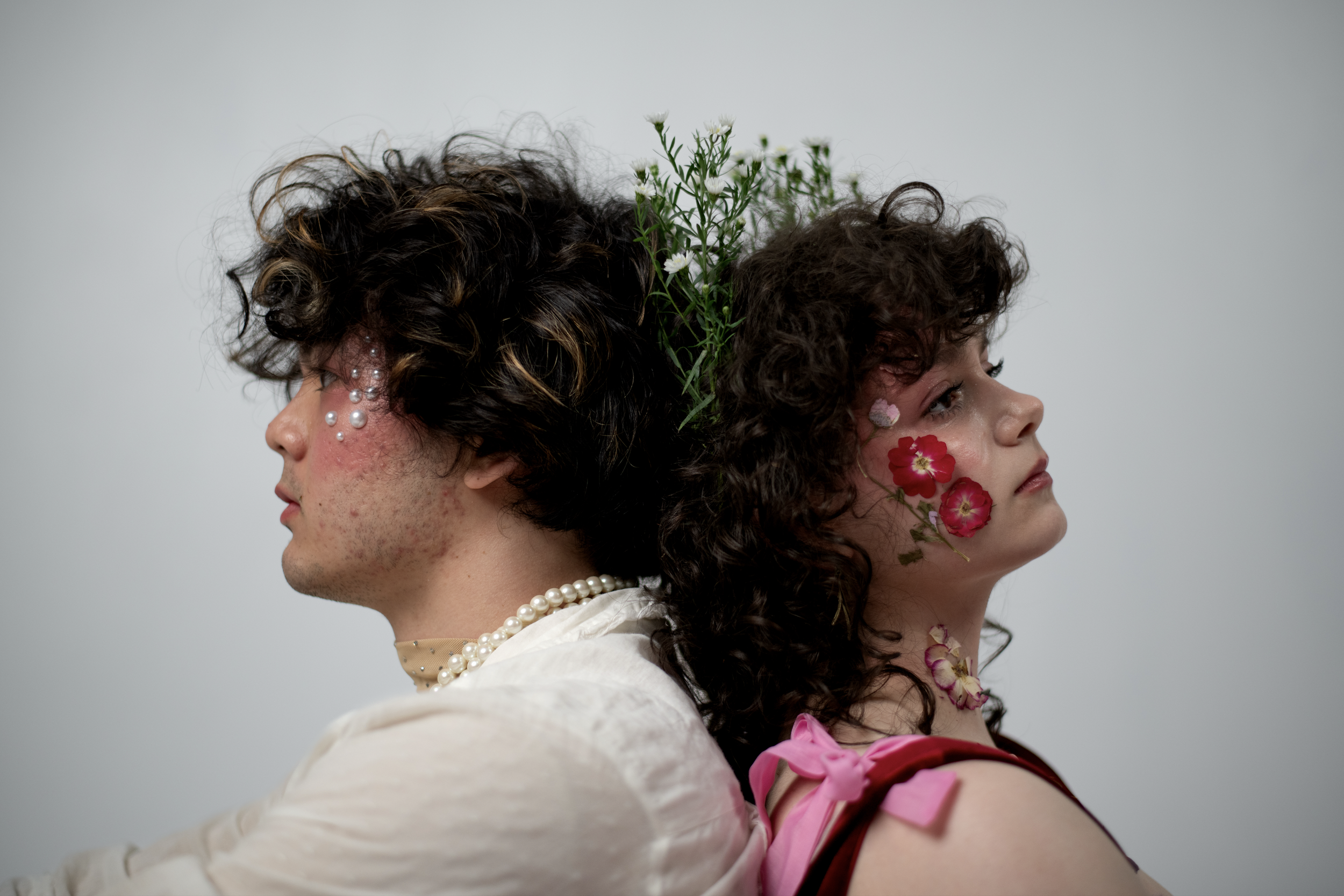Two individuals with curly hair and floral face decorations, standing back to back against a plain background. The person on the left with pearl embellishments and a pearl necklace, wearing a beige outfit, facing left. The person on the right with pink flowers on her face, wearing a pink outfit with a ribbon, facing right. Both have closed eyes and serious expressions, with flowers and greenery on their heads.