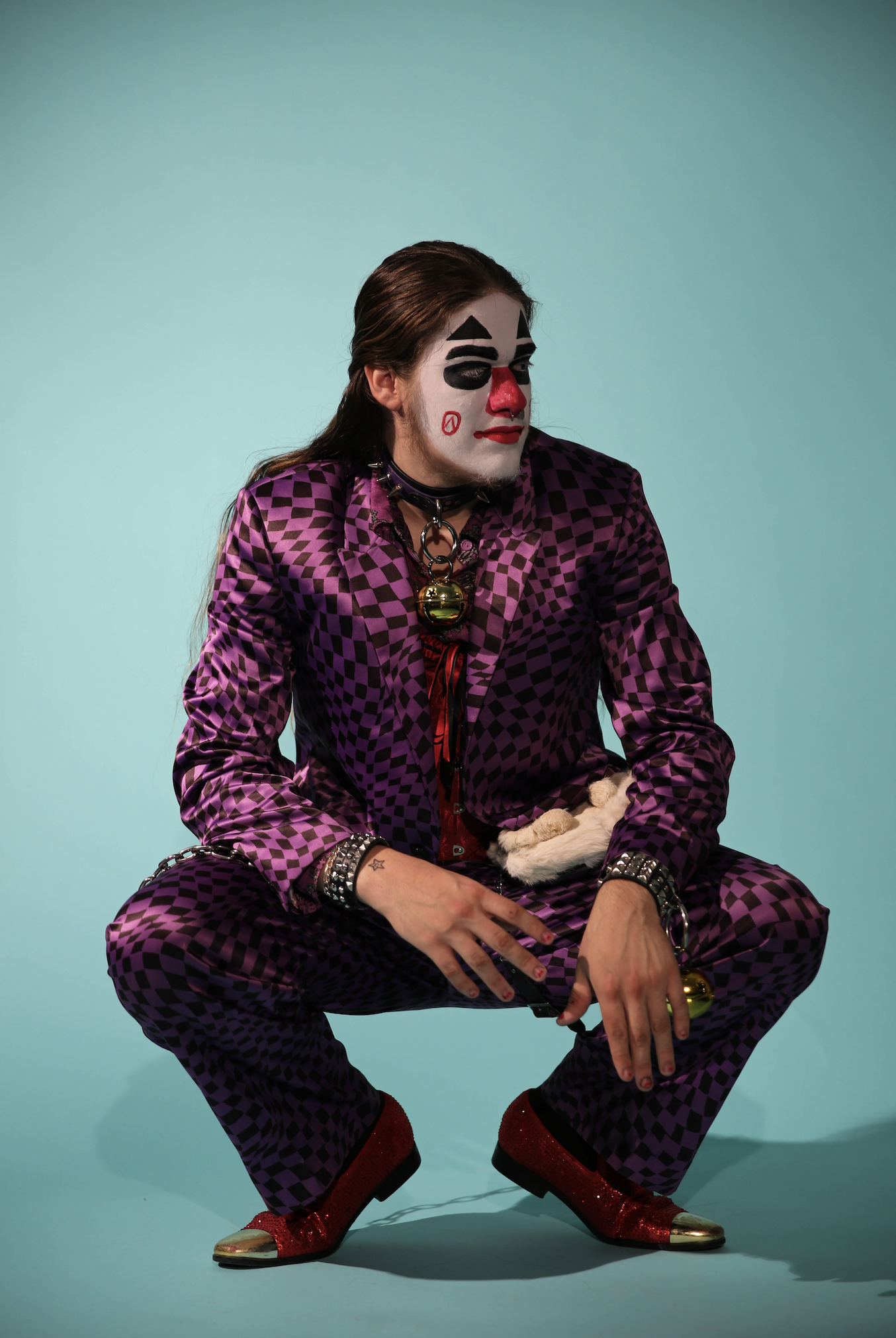 A person dressed as a clown with face paint, wearing a purple checkered suit, red glittery shoes, and various accessories, sitting against a light turquoise background.