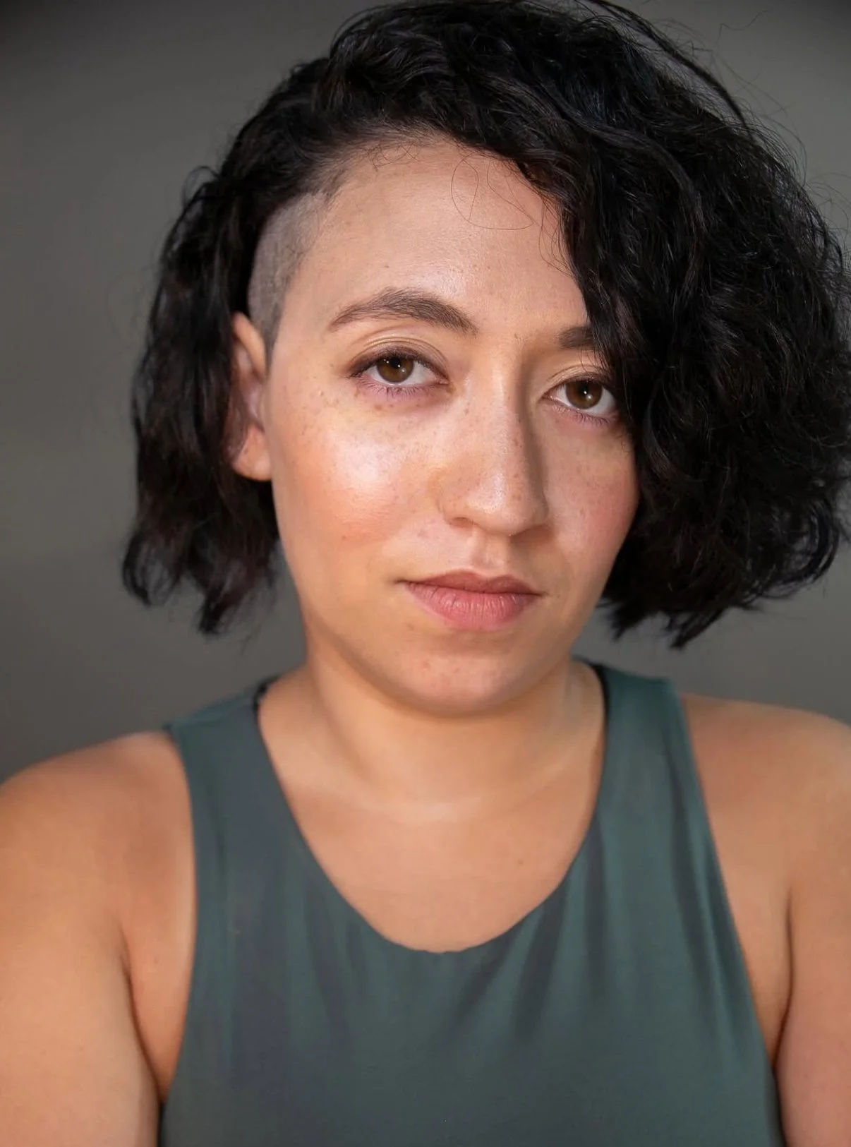 Close-up of a woman with short, curly, black hair, wearing a dark green sleeveless top, looking at the camera against a gray background.