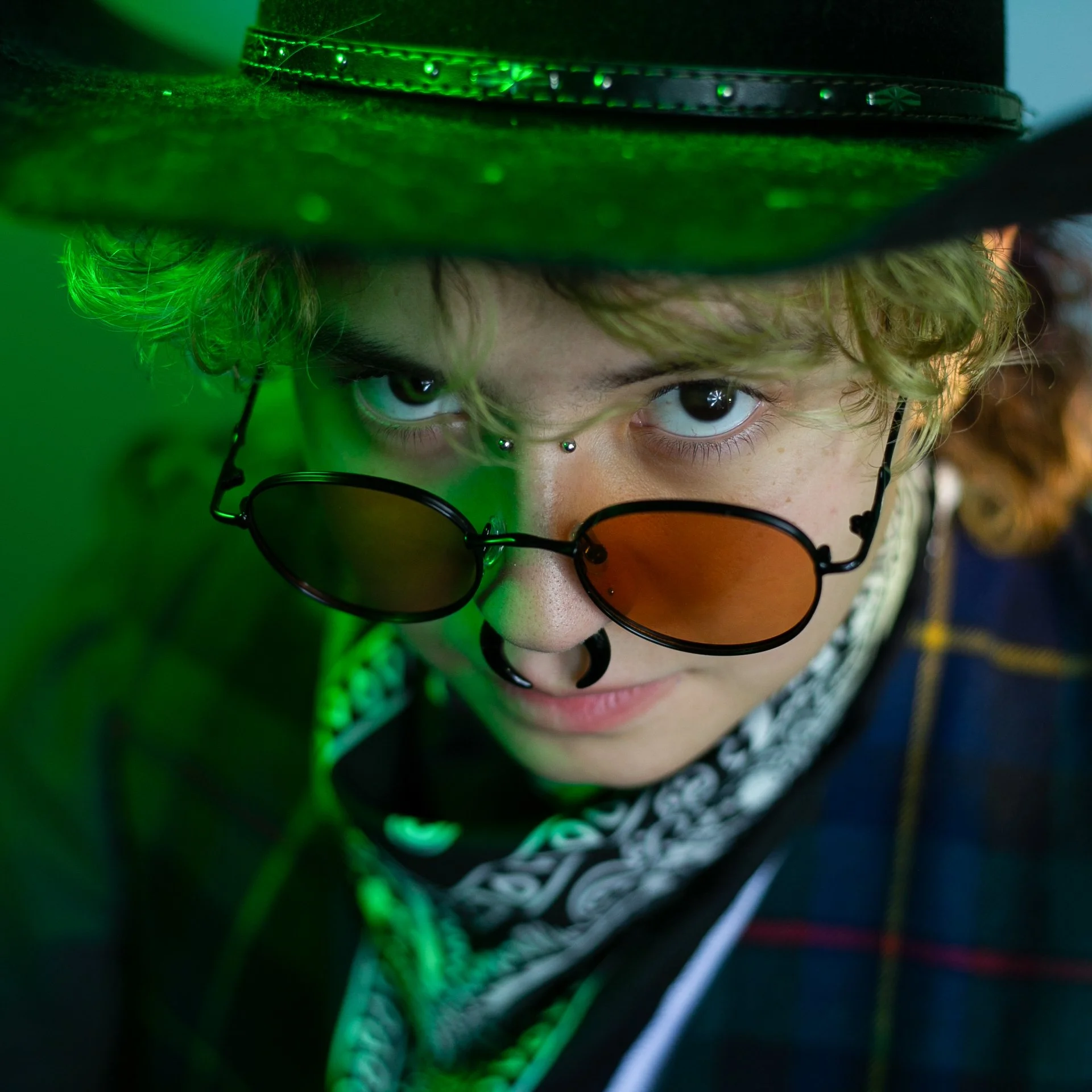 Close-up of a person wearing a green hat, orange-tinted sunglasses, a bandana, and a nose ring, with green lighting accenting their face and accessories.