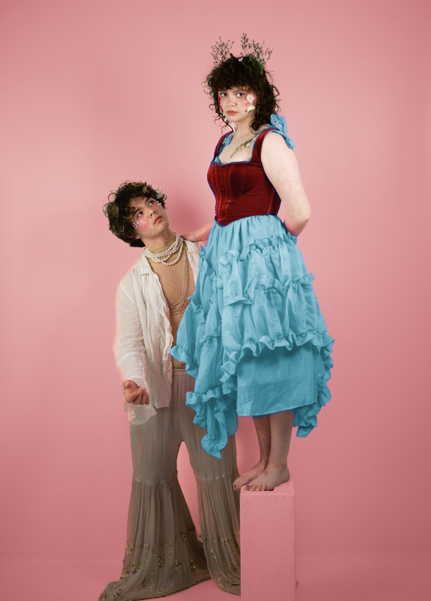 Two individuals dressed in colorful, elaborate costumes stand against a pink background. The taller person is standing on a pink platform, wearing a blue dress with a red velvet bodice and a floral crown, looking serious. The shorter person, kneeling and looking up at the taller individual, is dressed in a flowing beige outfit with pearl necklaces, makeup with glitter and face paint, and has curly hair.