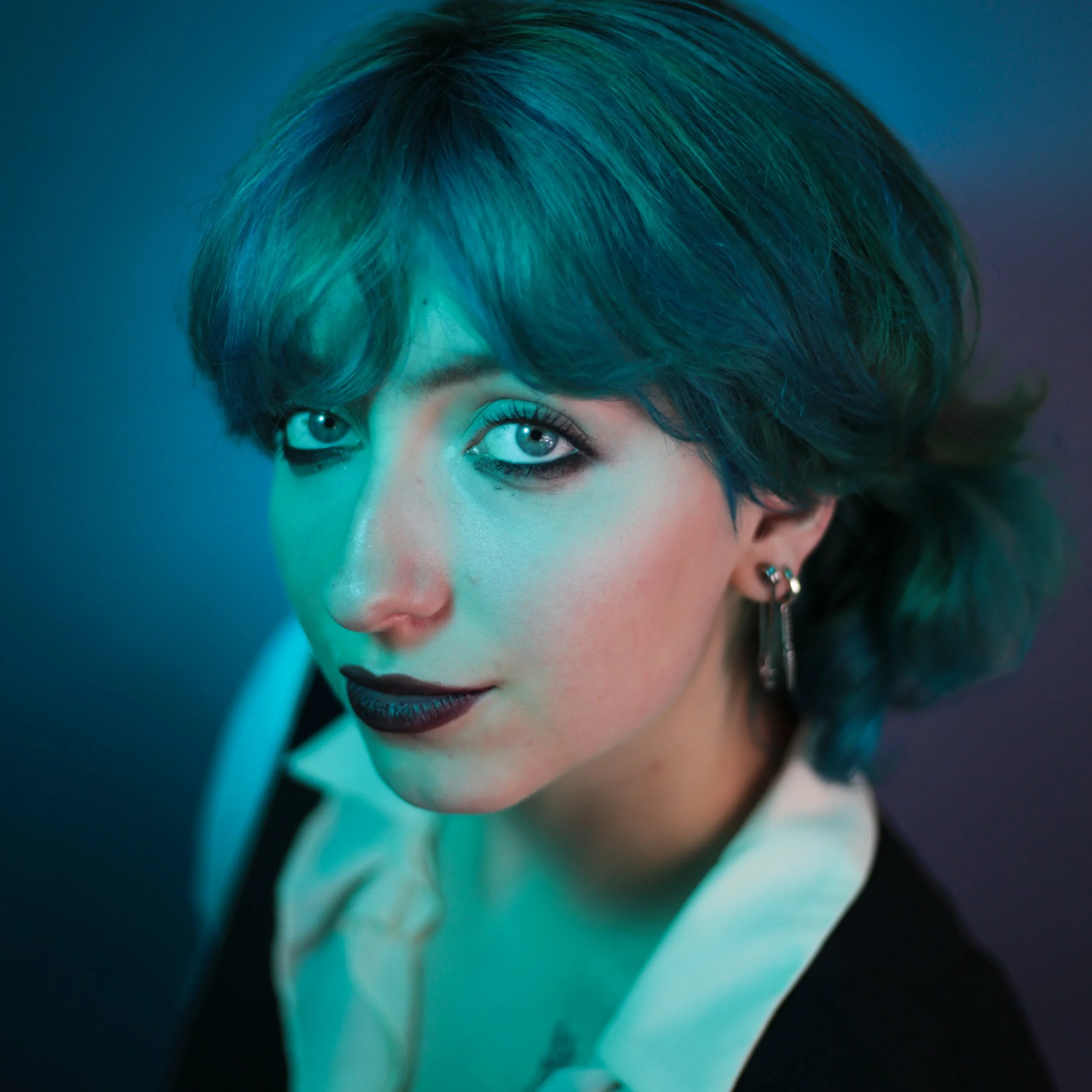A woman with short, edgy hair dyed in shades of blue and green, wearing dark lipstick, eyeliner, and earrings, looking directly at the camera with a confident expression.