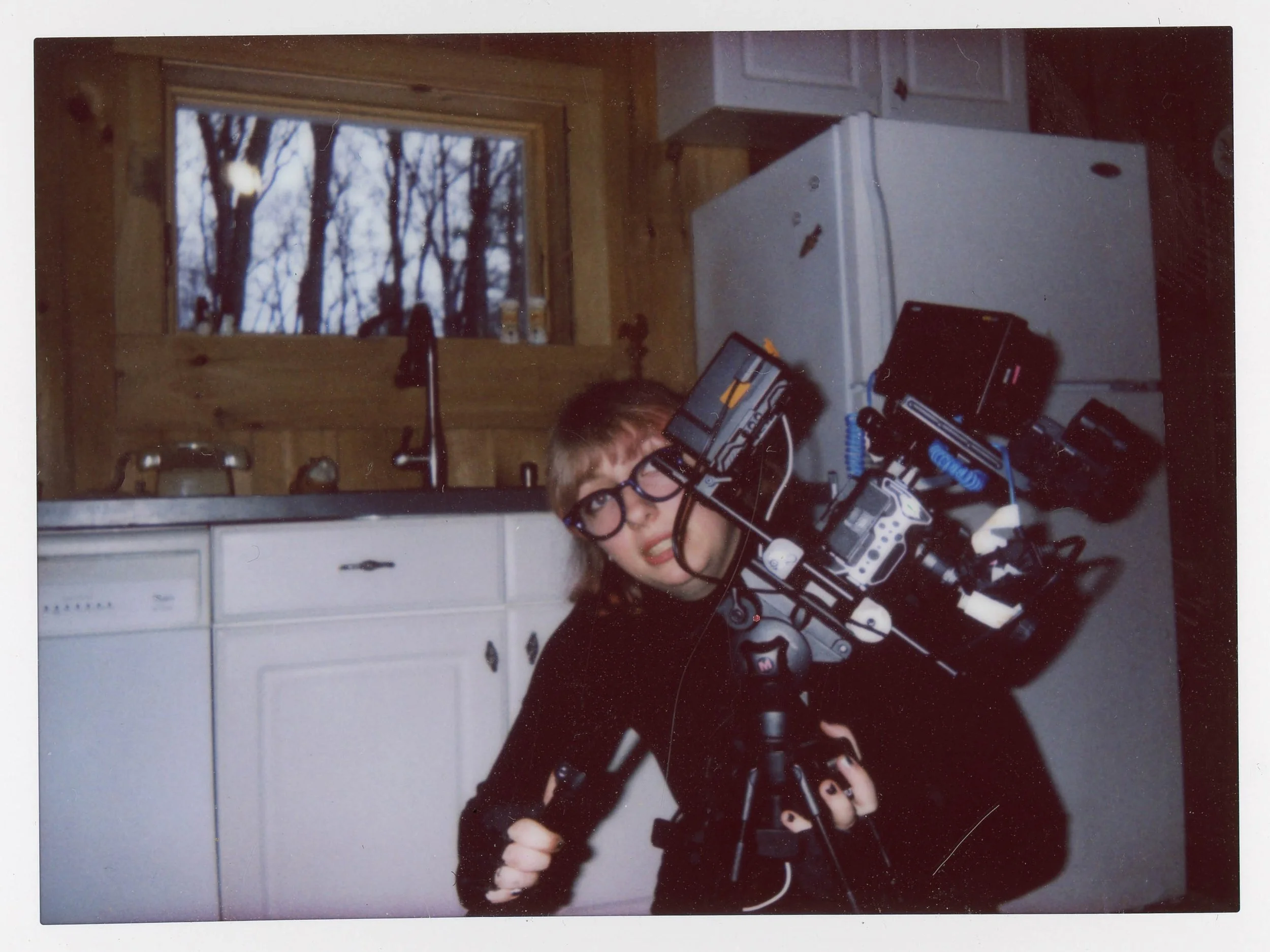 A woman with glasses holding a large professional camera on a tripod in a kitchen, with a window showing trees outside