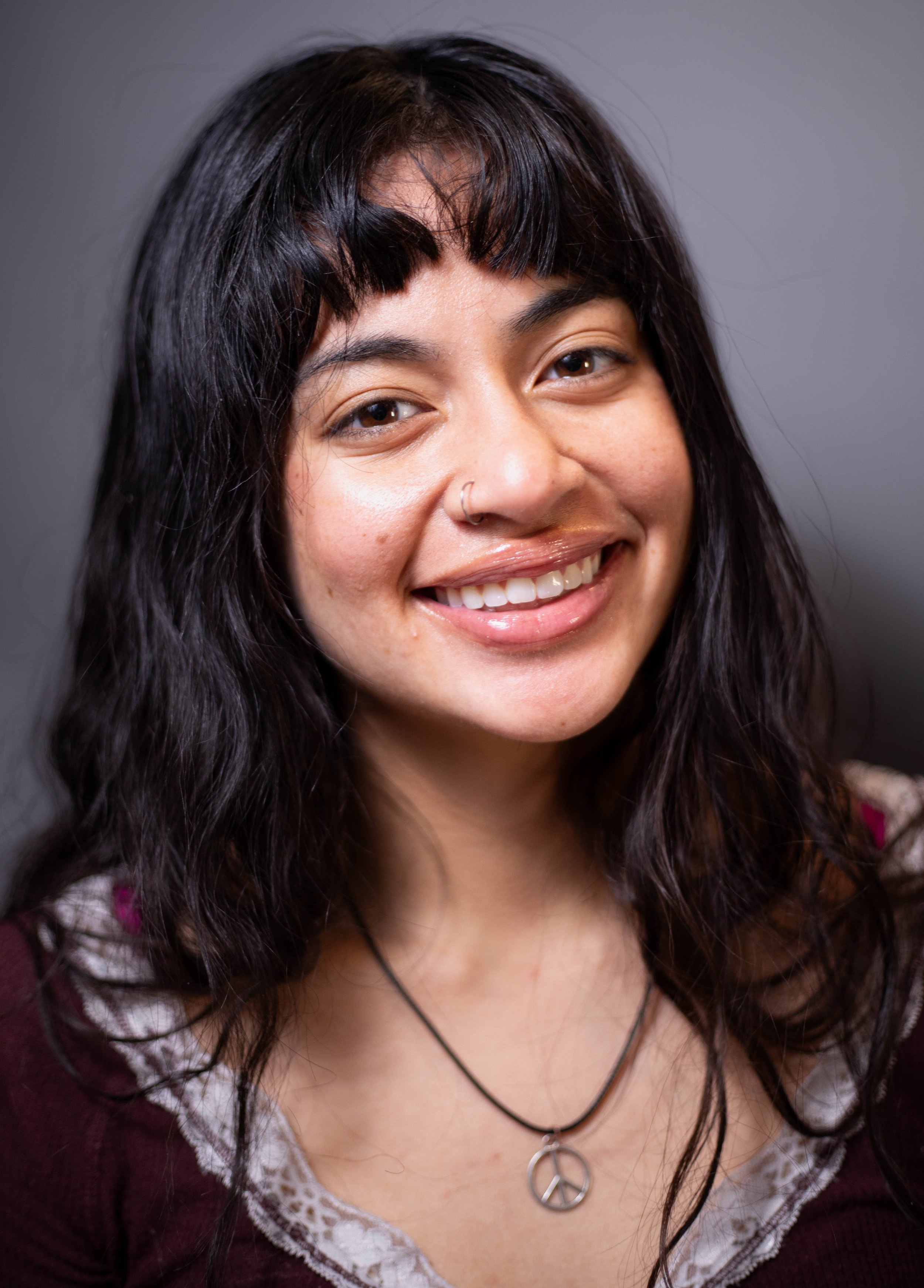 Close-up of a smiling woman with dark hair, wearing a peace sign necklace and a dark top, against a gray background.