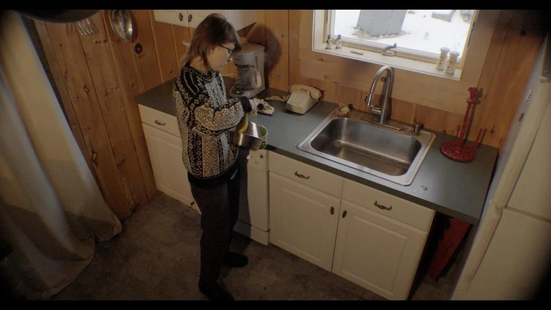 A person standing in a kitchen, pouring a liquid from a metal measuring cup into a bowl. The kitchen has wooden walls, white cabinets, a stainless steel sink, and a window above the sink.