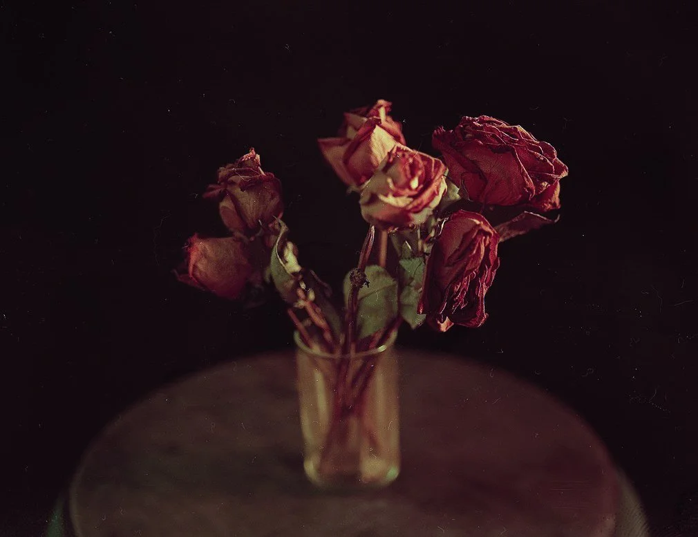 Dried red roses in a glass vase on a round table against a dark background.