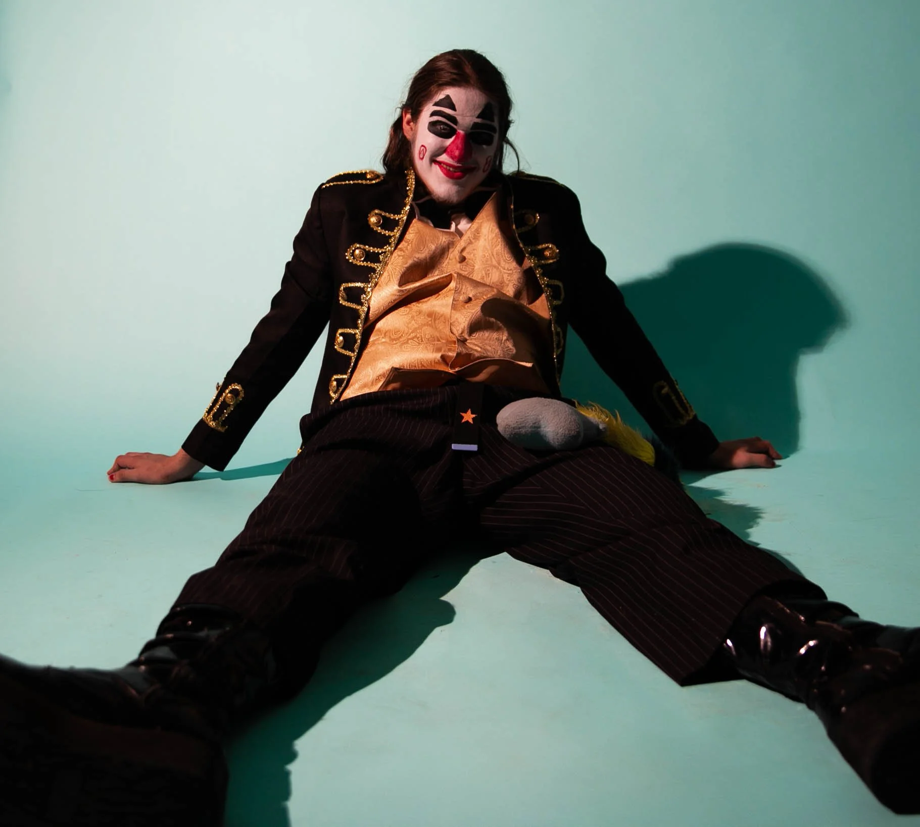 Person dressed as a clown with clown makeup, sitting on the floor with legs spread, wearing a black and gold costume and shiny black boots against a teal background.