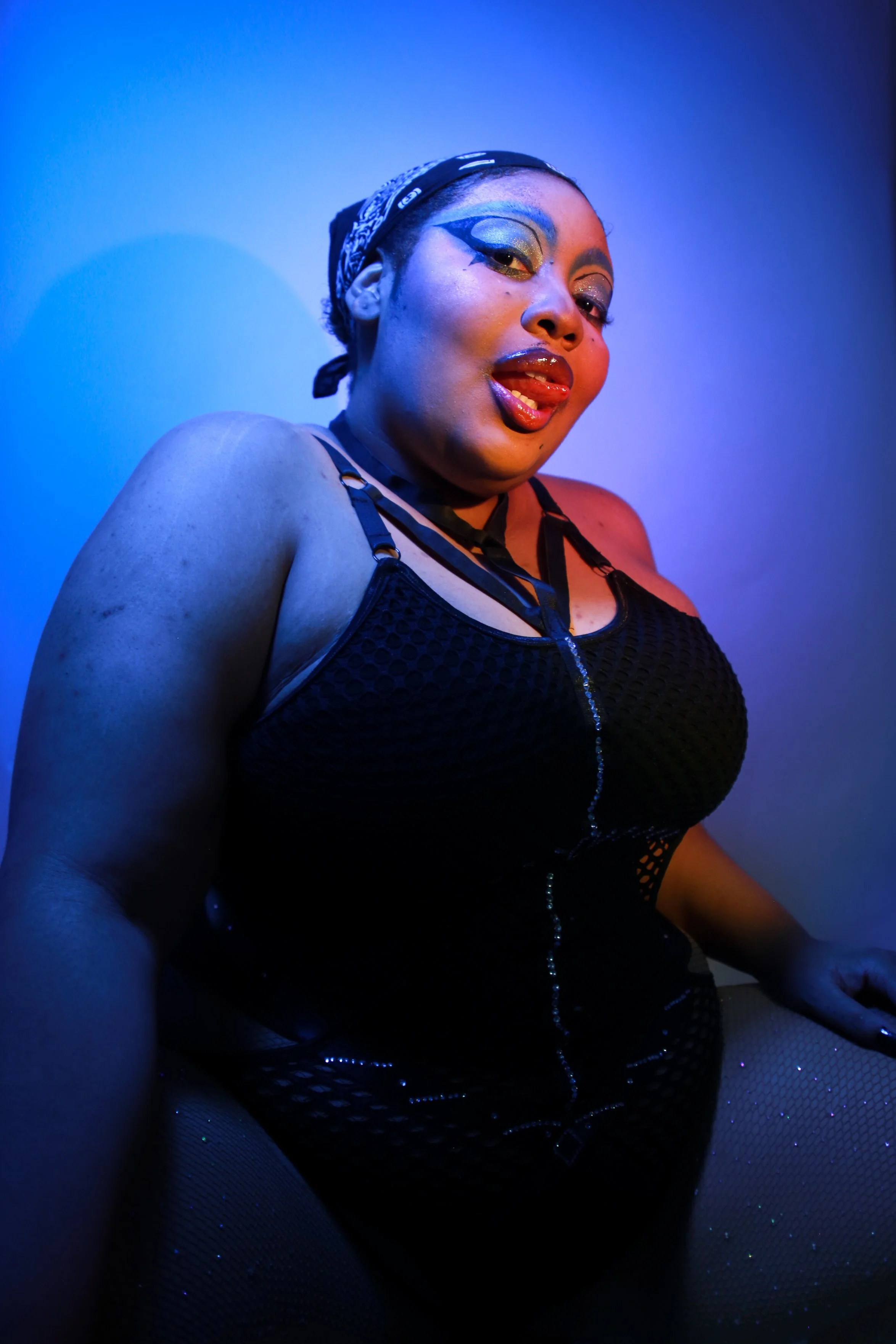 A woman with bold makeup, black mesh clothing, and a bandana on her head, posing under blue lighting.