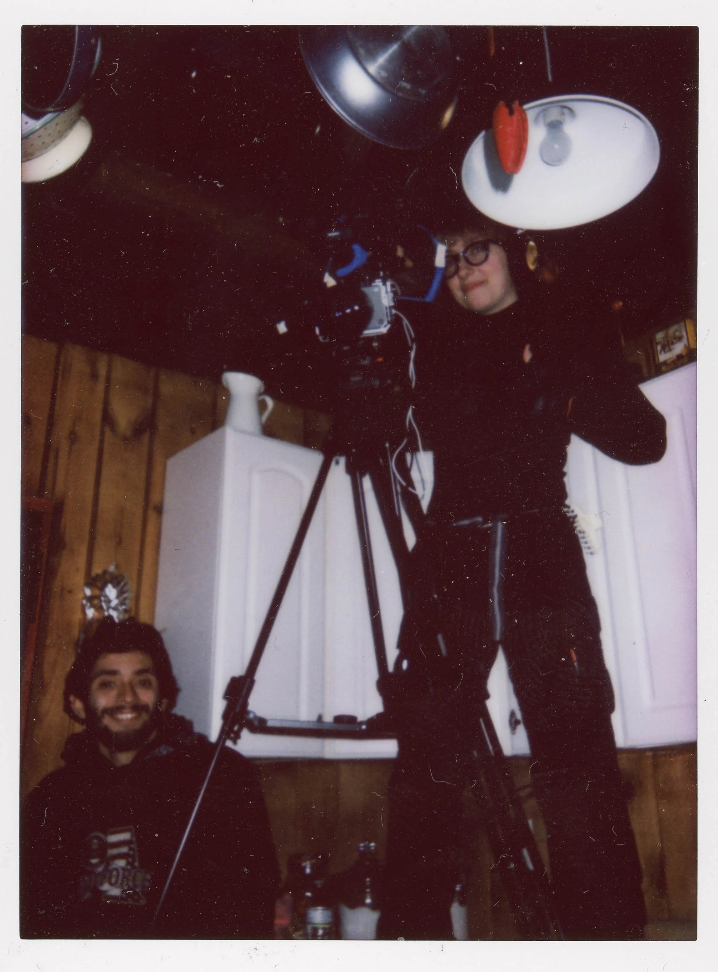 Two people with a camera on a tripod inside a room with wooden walls. One person is sitting with a smile, and the other is standing on a chair, adjusting the camera, looking at the camera, wearing glasses and a black outfit. The ceiling is decorated with hanging pots and pans, and there are various bottles on the floor.