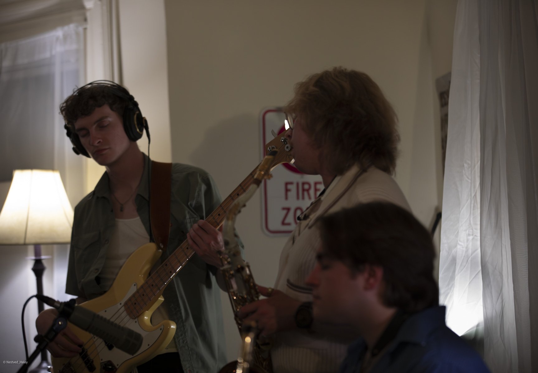 Three young men playing musical instruments indoors, with curtains and a lamp in the background.
