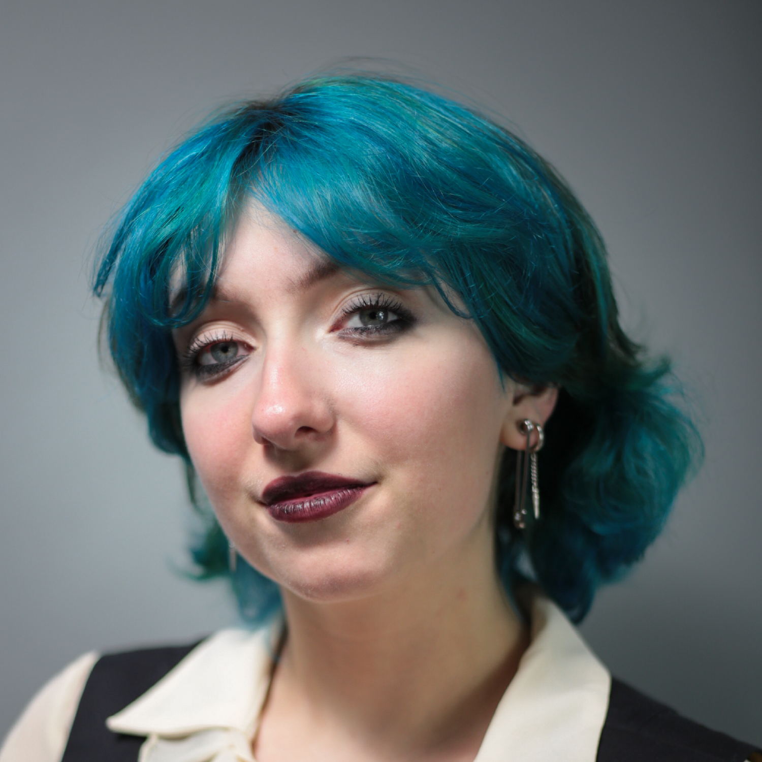 A person with short, wavy blue hair, wearing earrings, a light-colored blouse, and a dark vest, looking at the camera with a slight smile against a neutral background.