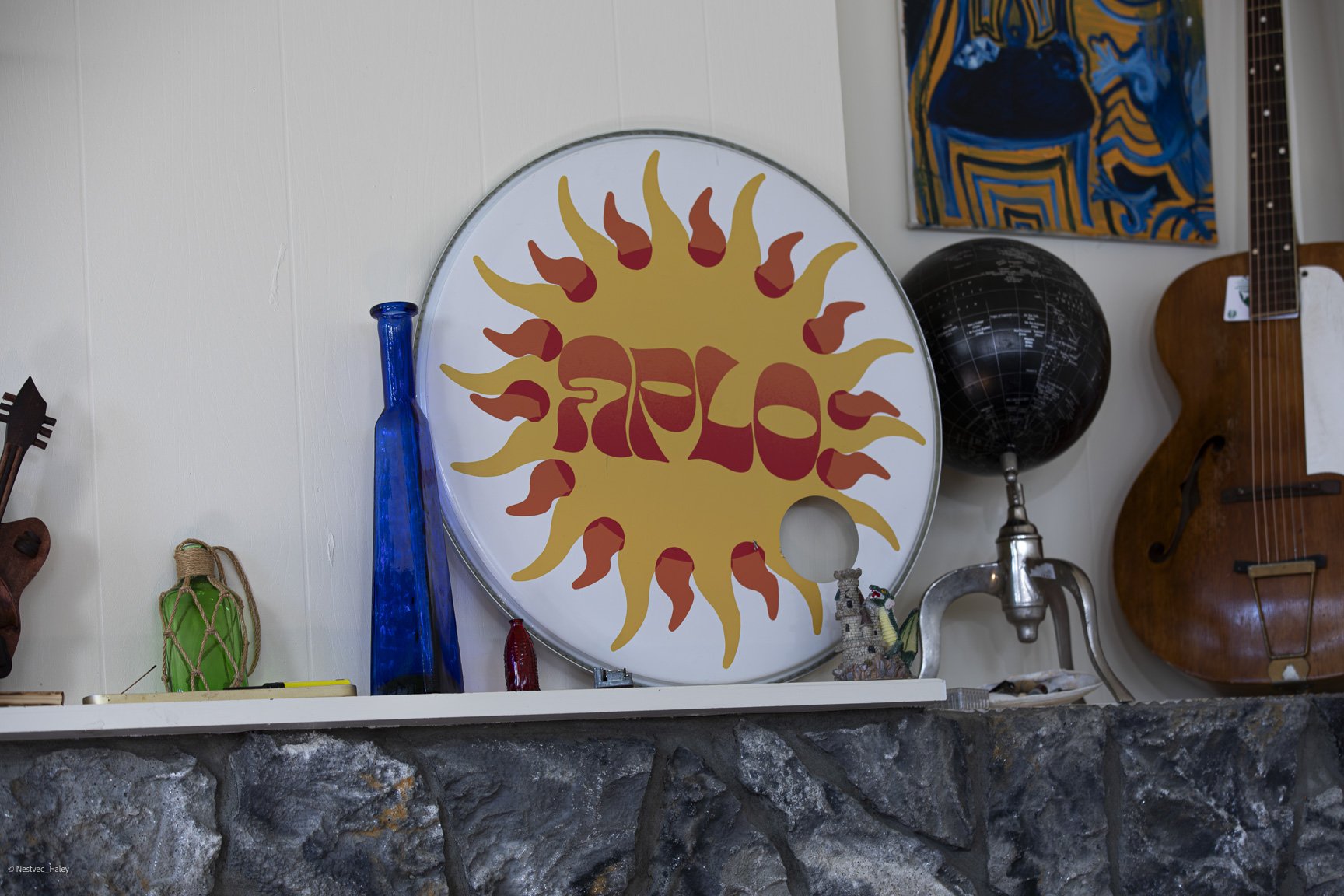 Decorative wall shelf with various objects including a large circular sign with a sun design and the word 'FUEL' in the center, two tall blue glass bottles, a small red bottle, a small green lantern, a painted canvas, a black globe, a wooden sculpture, and part of a wooden guitar.
