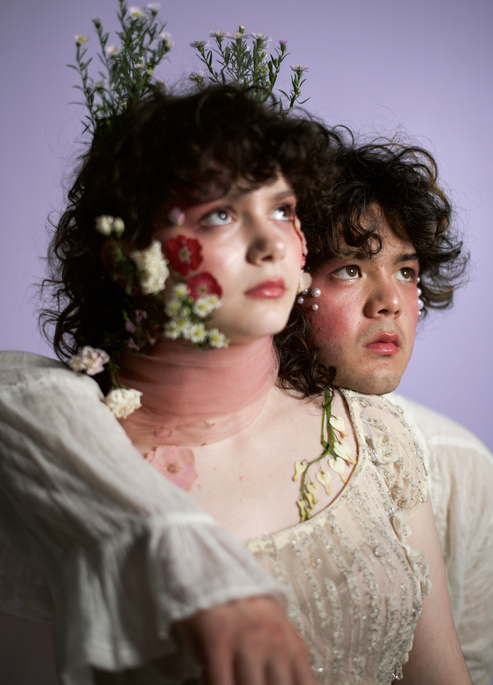 Two young people with floral makeup and accessories, facing upward, against a purple background.