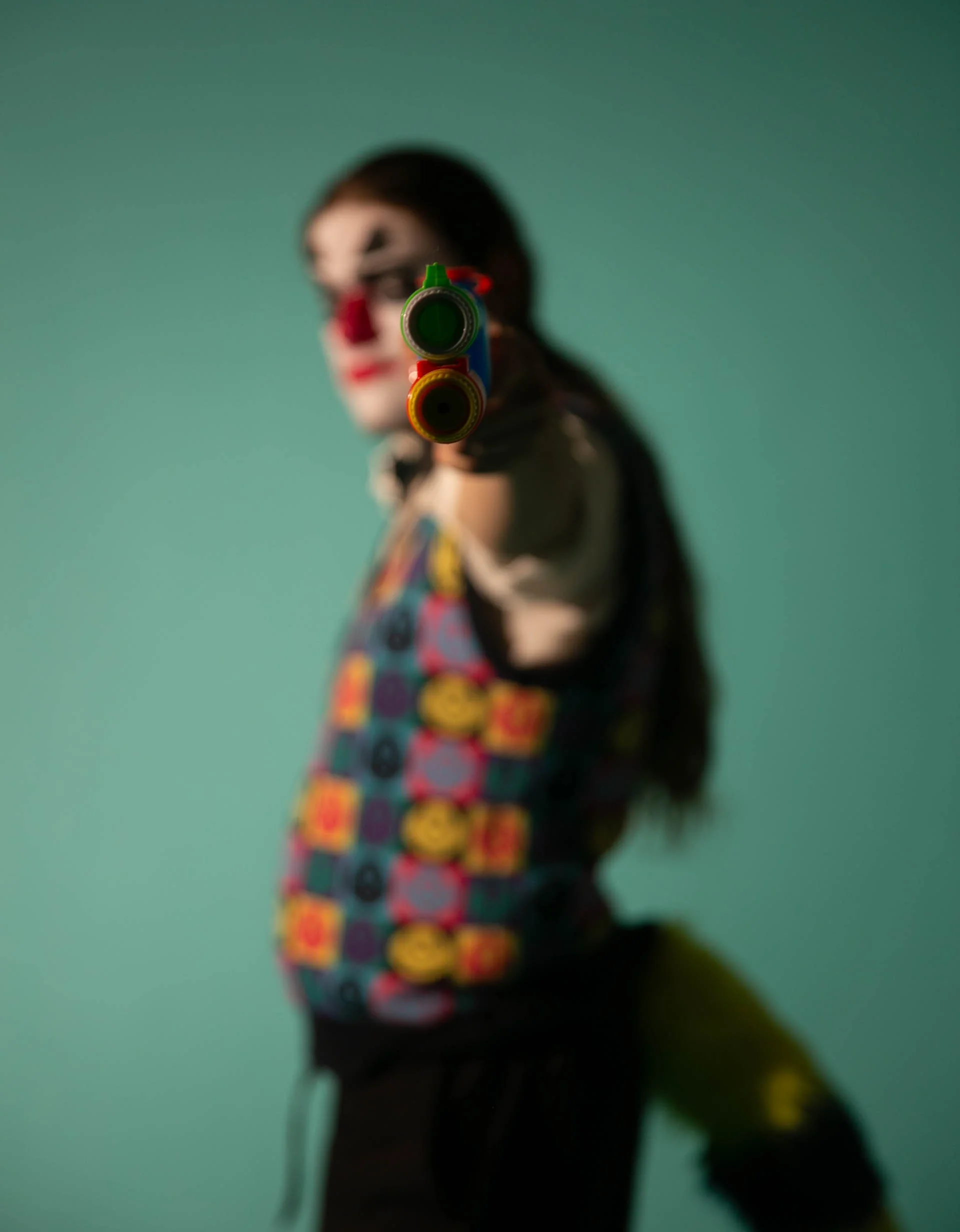 A person dressed as a clown with a painted face, wearing a colorful vest with smiley face patterns, pointing a toy gun directly at the camera.