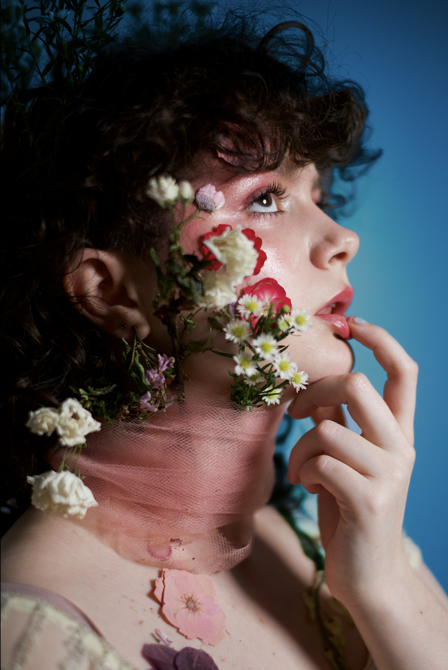 A person with curly hair and makeup, adorned with flowers on his face, neck, and shoulder, touching their lips with his finger, against a blue background.