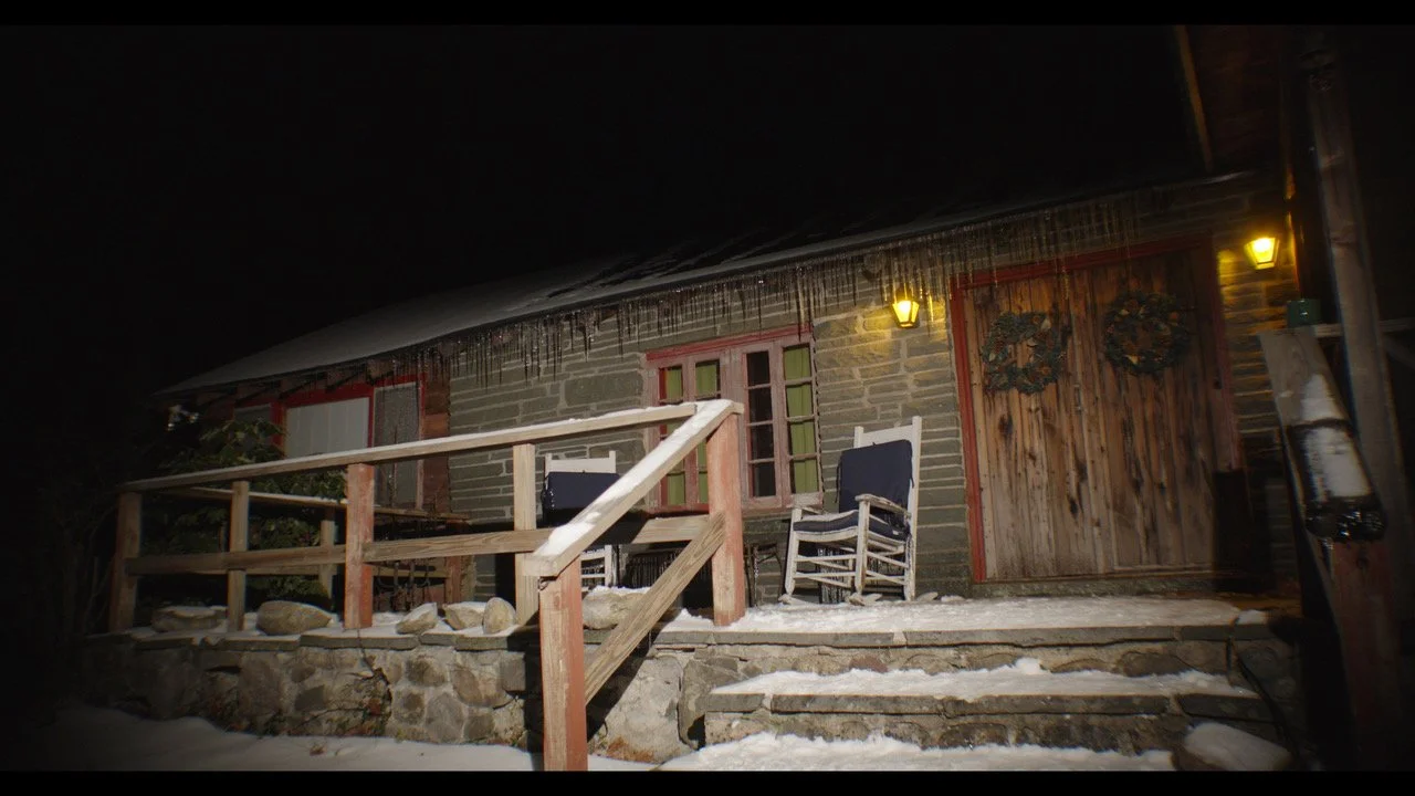 A rustic wooden cabin with stone foundation, two rocking chairs, Christmas wreaths on the door, and icicles hanging from the roof during snow at night, lit by yellow exterior lights.