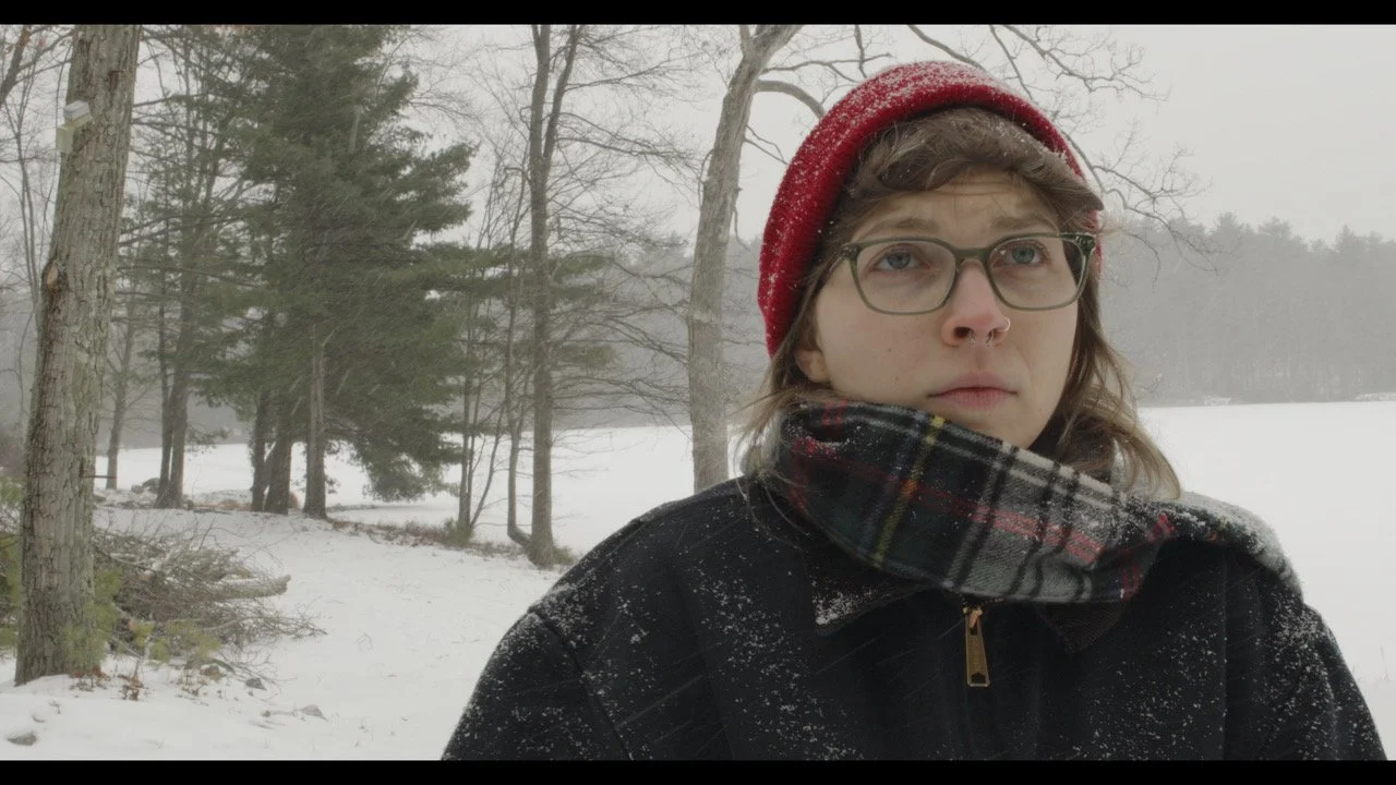 A person with glasses, wearing a red knit hat, a plaid scarf, and a dark winter jacket, standing outdoors in a snowy landscape with trees in the background.