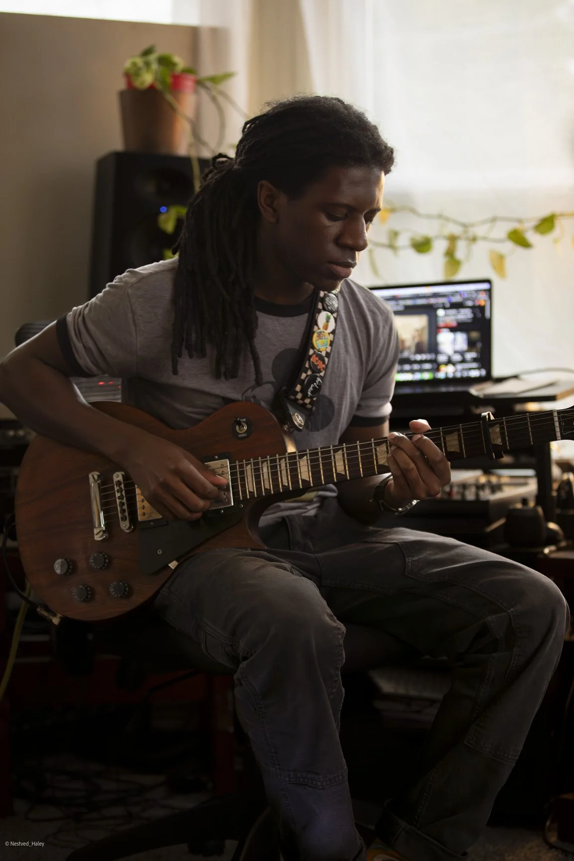 A young man with dreadlocks sitting and playing an electric guitar in a music studio with equipment and a computer in the background.
