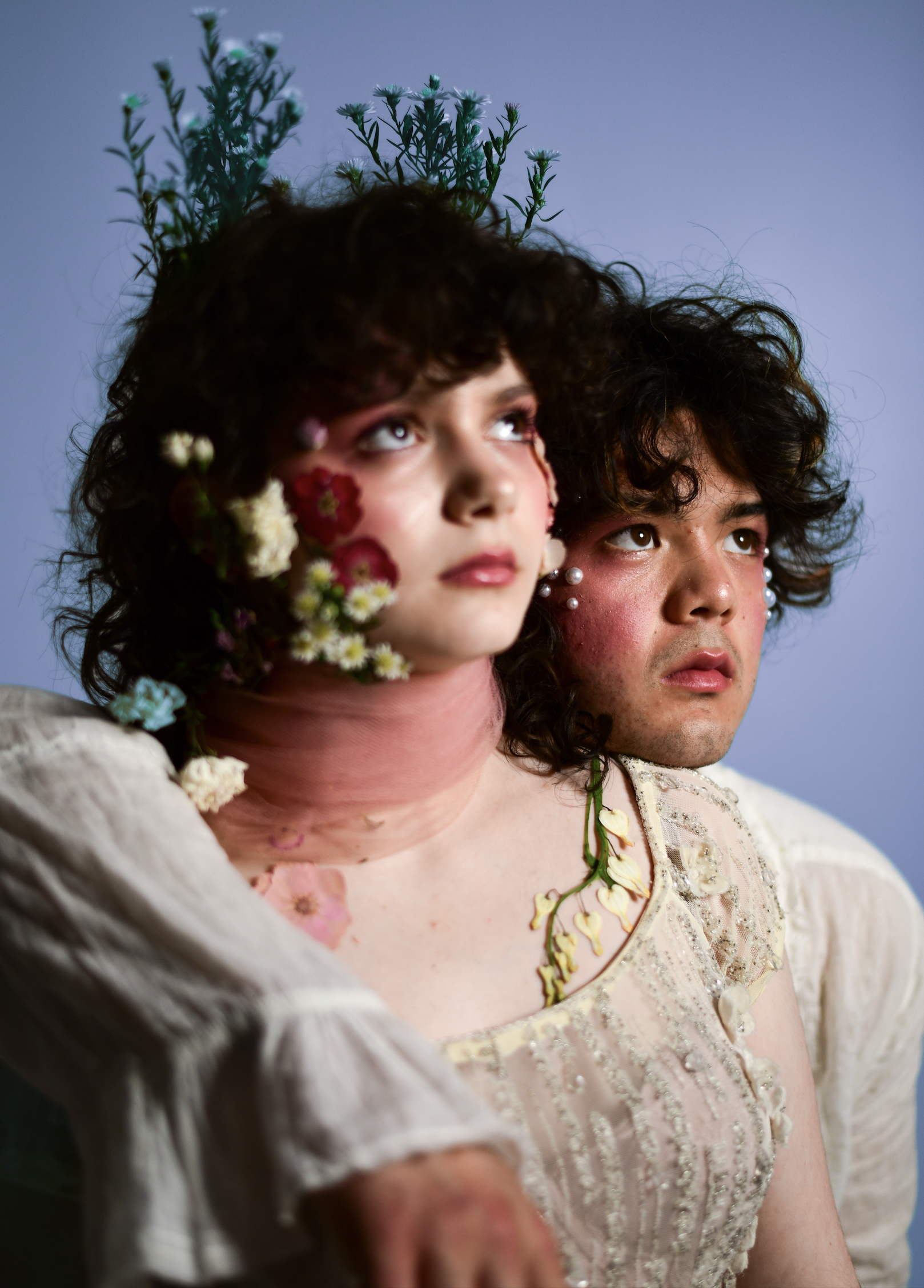 Two people, one with flowers and foliage in her hair, and makeup resembling flower petals on her face, and the other with makeup and earrings, both looking upward against a blue background.