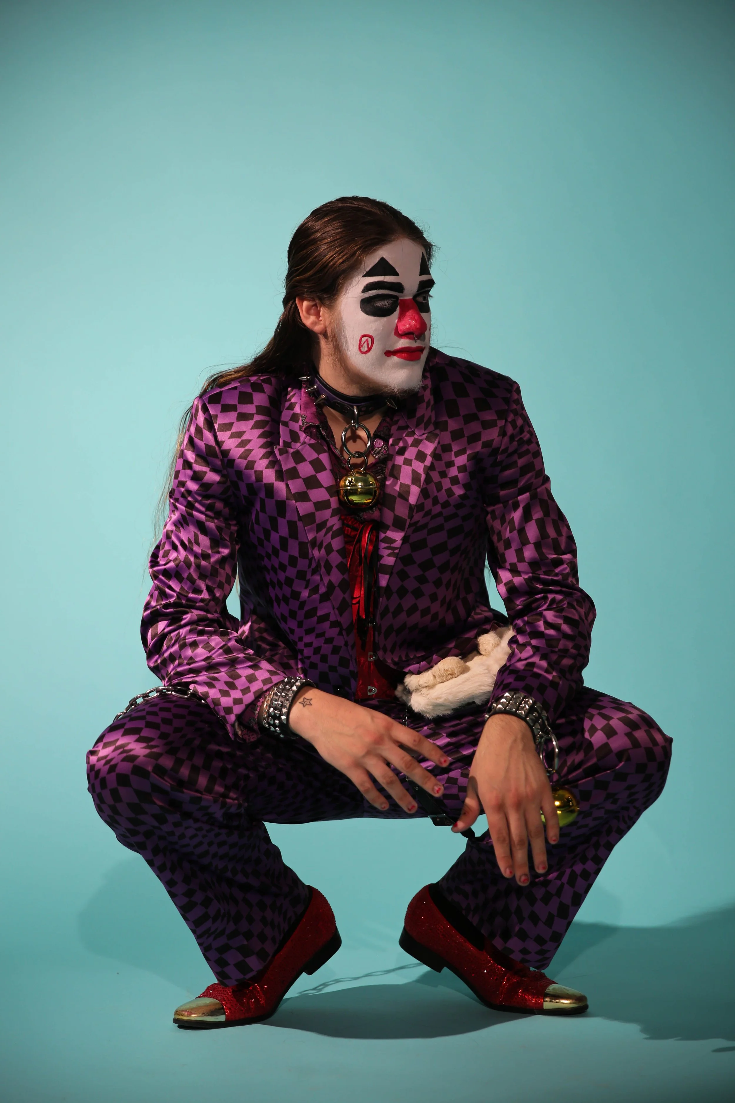 Person dressed as a clown with clown makeup, purple checkered suit, and red glitter shoes, squatting against a teal background.