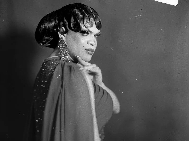 Black and white photograph of a woman with styled hair, wearing large earrings and elegant clothing, posing with her hand near her chin against a plain background.