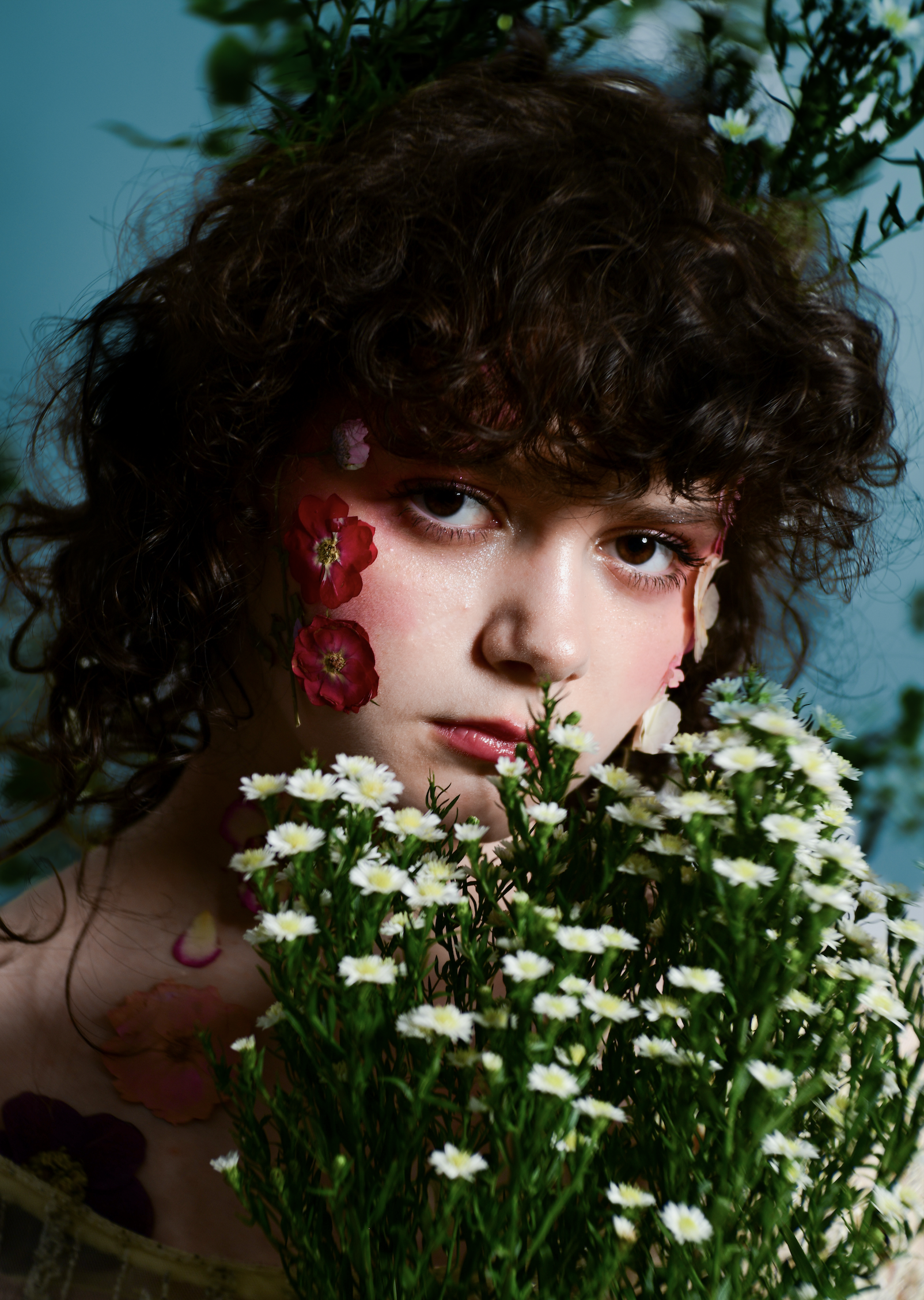 A person with curly brown hair and light makeup looks directly at the camera surrounded by flowers. he has red, pink, and white flower petals on his face and flower petals on her shoulders, with a bunch of white daisies in front of him.
