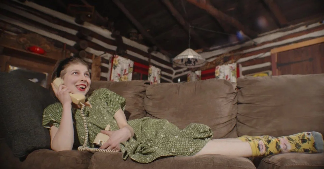 A woman with dark hair lying on a brown couch, talking on a landline phone and smiling, in a rustic log cabin interior.