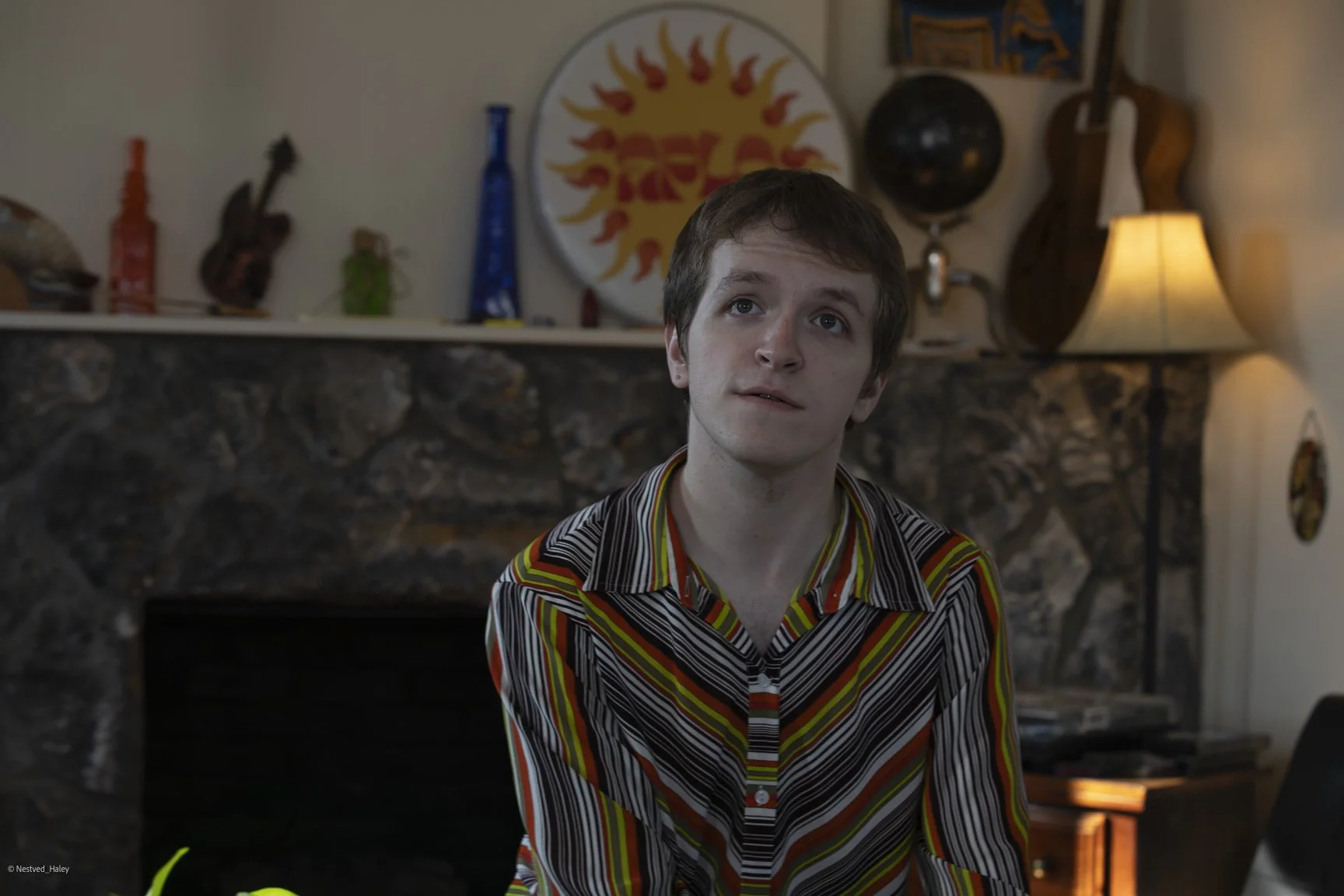 A young man with short brown hair and fair skin, wearing a multicolored striped shirt, sitting in a cozy living room with a stone fireplace in the background, decorated with various items including guitars, a bowl, and colorful artwork, with warm lig