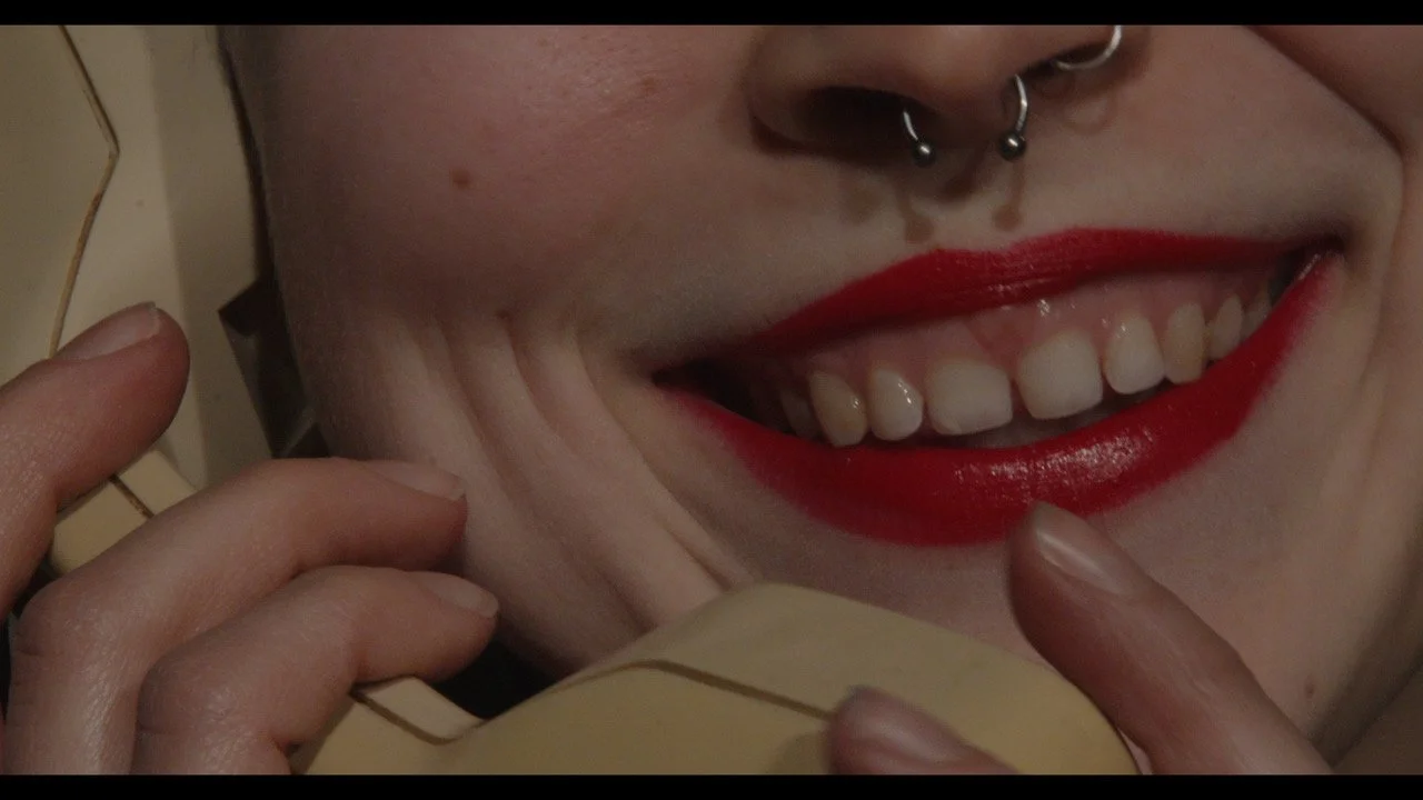 Close-up of a person smiling while talking on a vintage rotary phone, wearing bright red lipstick and some jewelry.