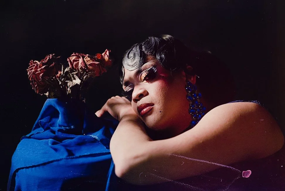 A woman with vintage style hair and makeup, wearing a blue dress and matching earrings, is lying down on a black background, resting her head on her arm near a bouquet of dried red roses.