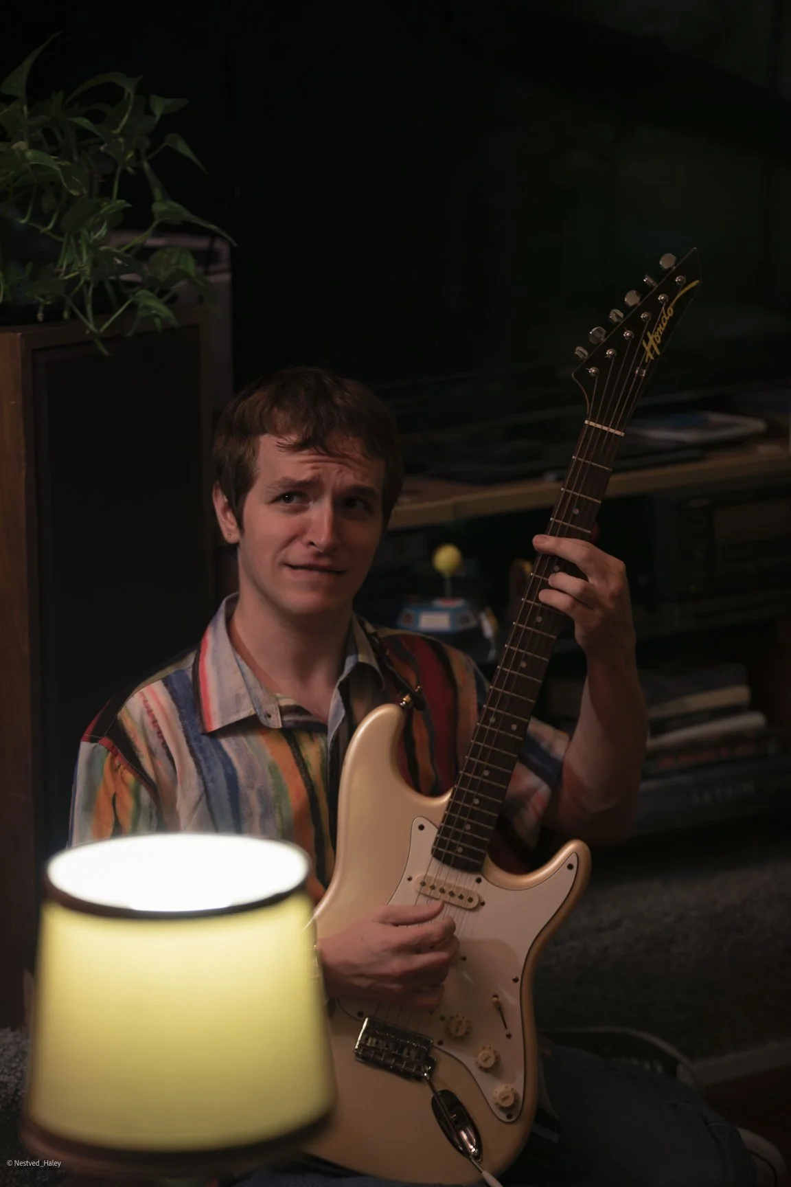 A young man with a puzzled or confused expression holding an electric guitar in a dimly lit room.