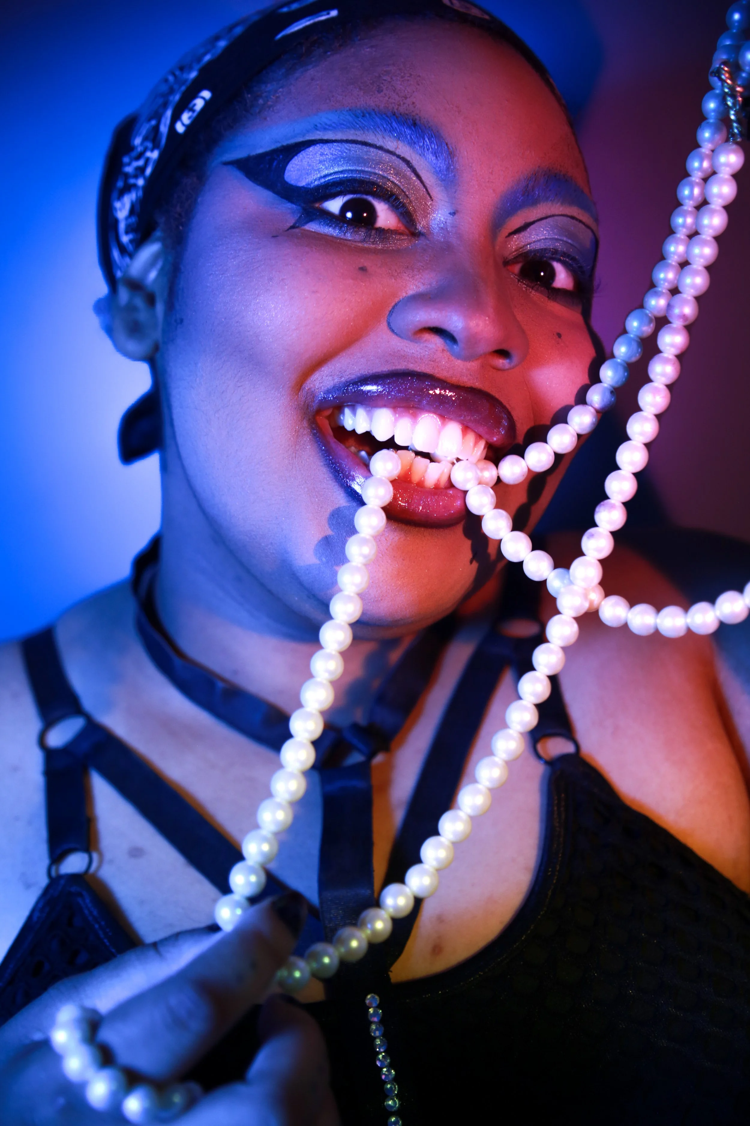 A person with bold, colorful makeup smiling, holding a string of pearls, wearing a black lace top and a black bandana, with dramatic lighting.