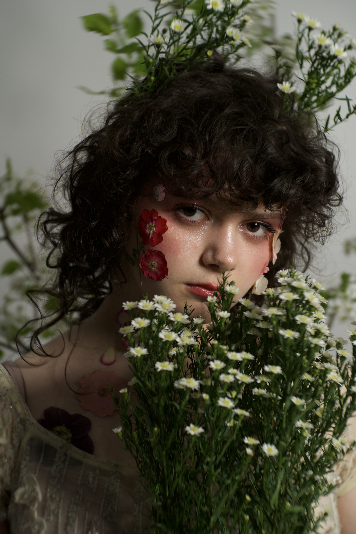 A young woman with curly hair surrounded by white and pink flowers, some on her face and neck, with additional flowers and greenery in the background.
