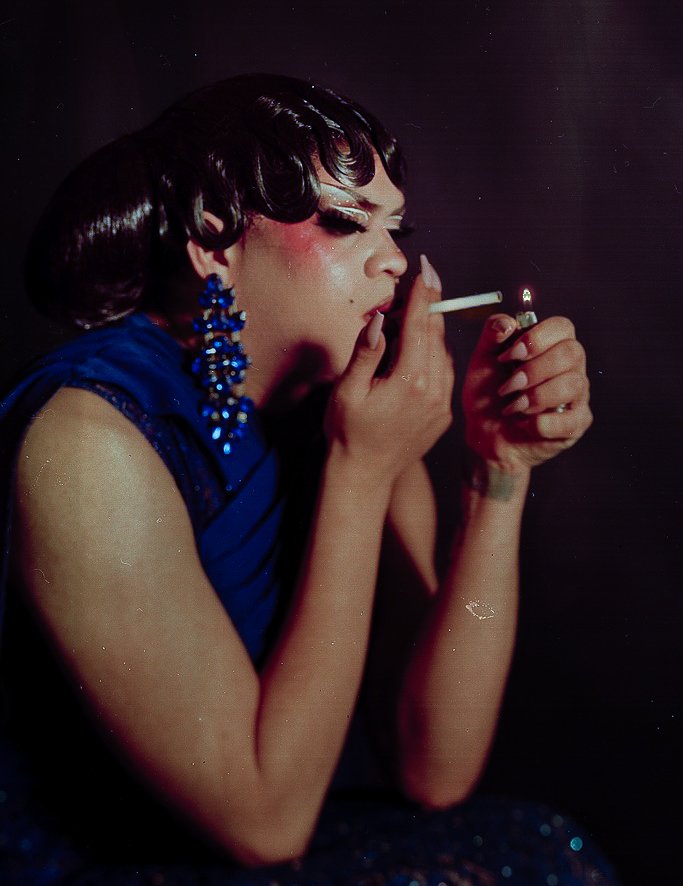 Person with dark hair and makeup, wearing blue earrings and dress, lighting a cigarette in a dark setting.