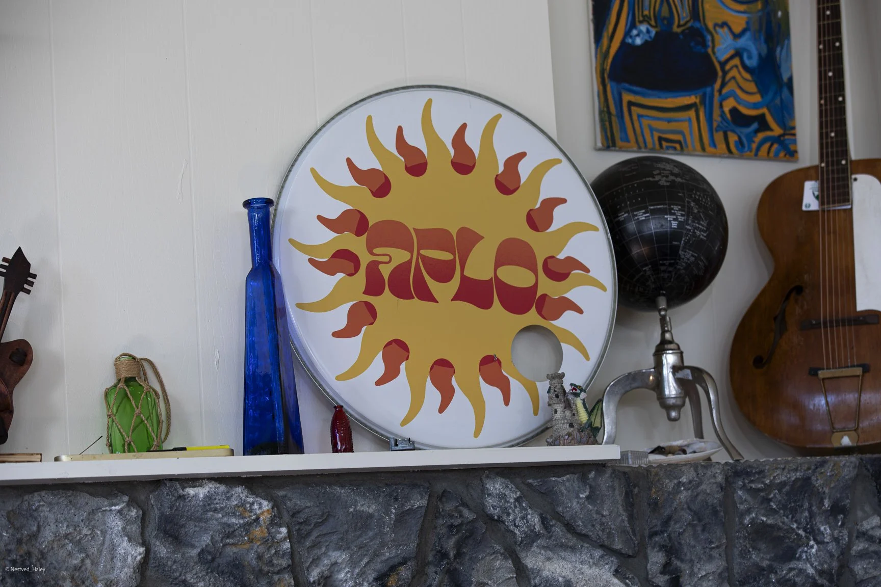 Decorative shelf with a round colorful sign featuring a sun design with red and yellow flames and the word 'SOL' in stylized red letters, along with glass bottles, a globe, a wooden guitar, and small decorative objects.